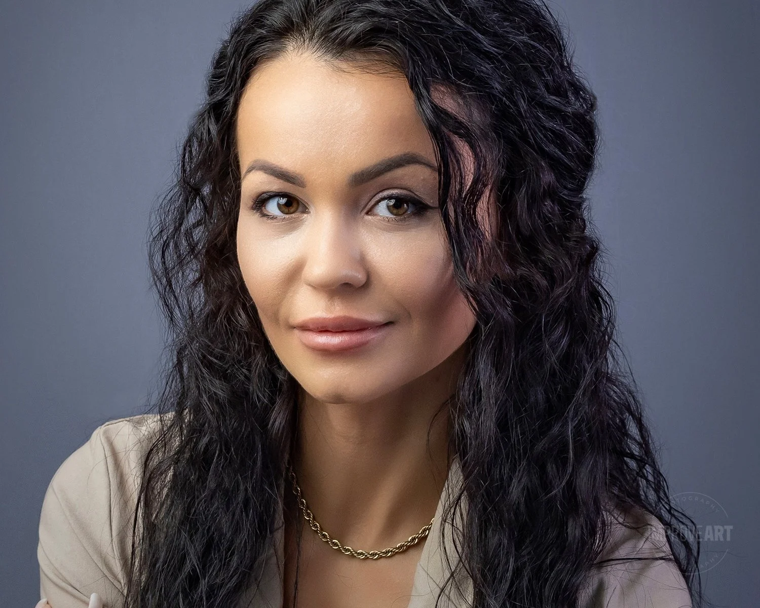 A woman with long, curly black hair, tan skin, and brown eyes, wearing a beige blazer and a gold necklace, looking slightly to the right with a soft smile, against a gray background.