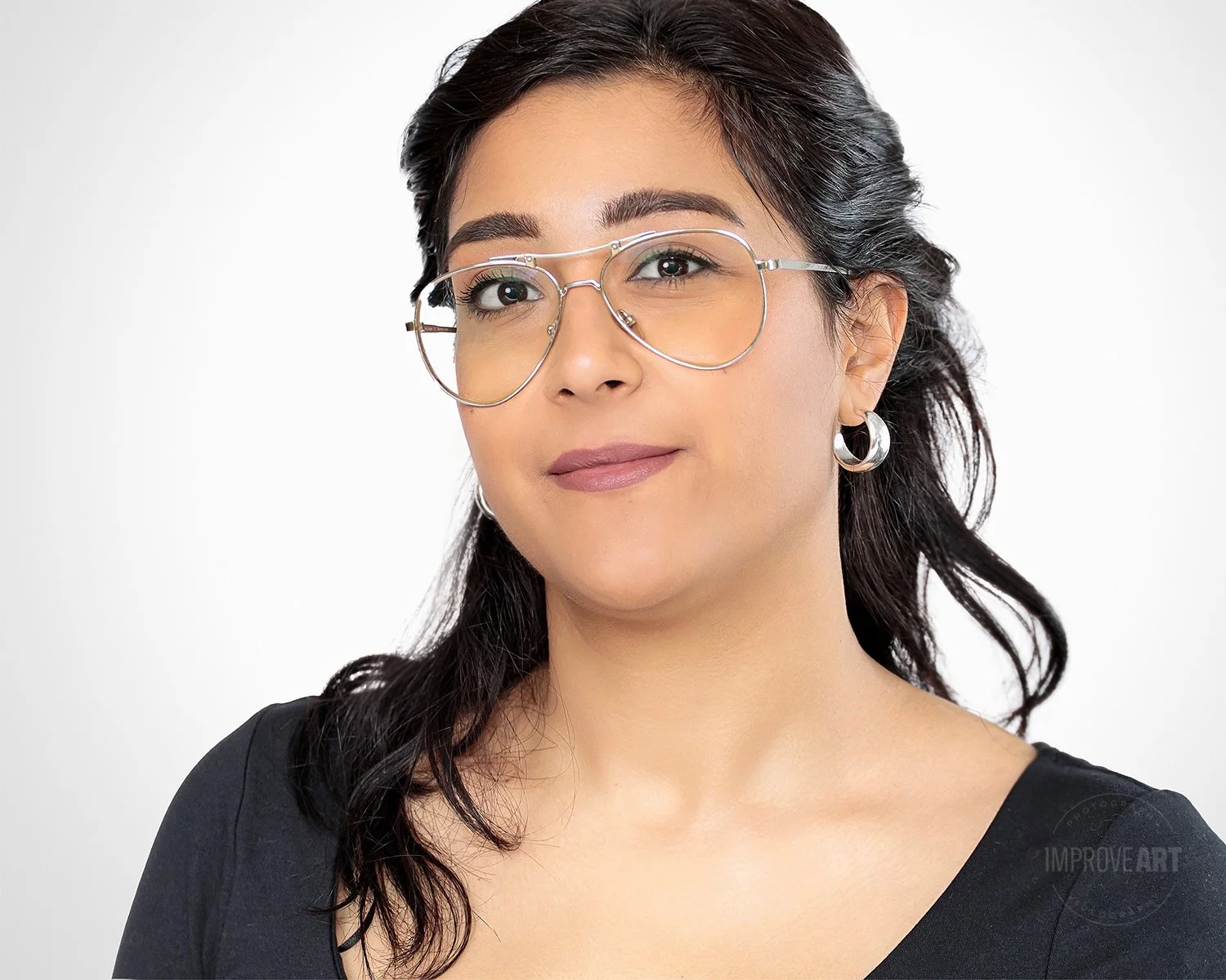 A woman with dark hair in loose waves, wearing silver hoop earrings and large silver-rimmed glasses, looking confidently at the camera against a plain light background.