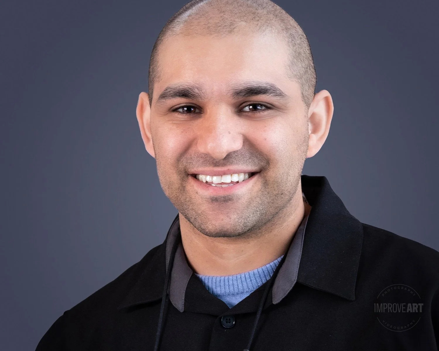 Headshot of a smiling man with a shaved head, wearing a black jacket over a blue shirt, against a gray background.
