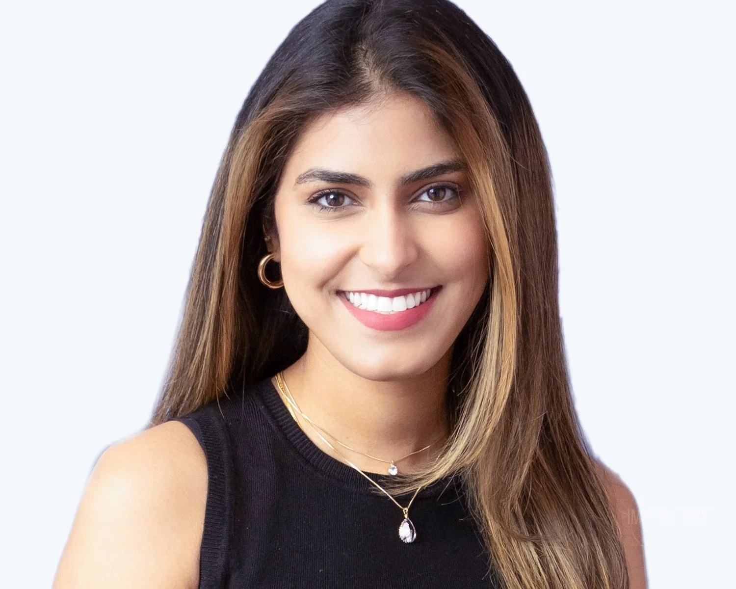 Close-up portrait of a smiling woman with long brown hair wearing a black sleeveless top, gold jewelry, and bright makeup, against a plain light background.