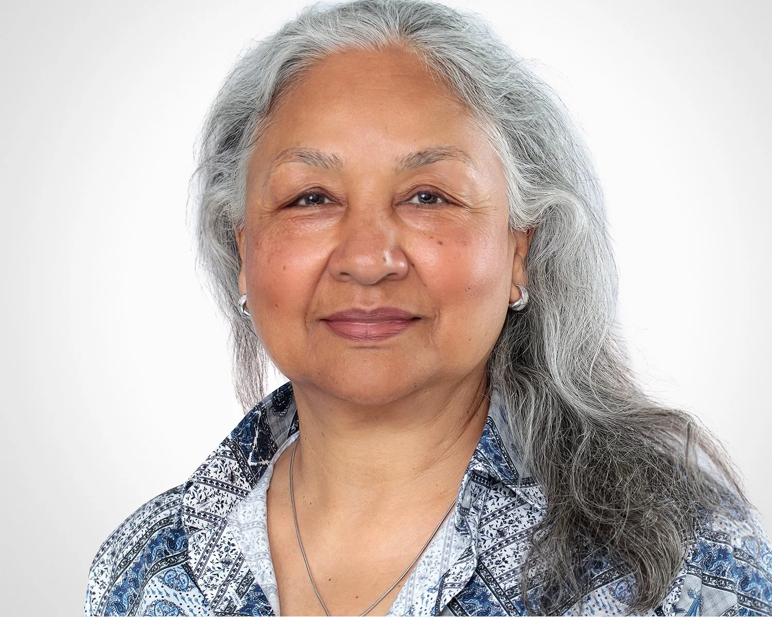 A middle-aged woman with long gray hair, wearing a patterned shirt and silver jewelry, smiling softly against a plain white background.