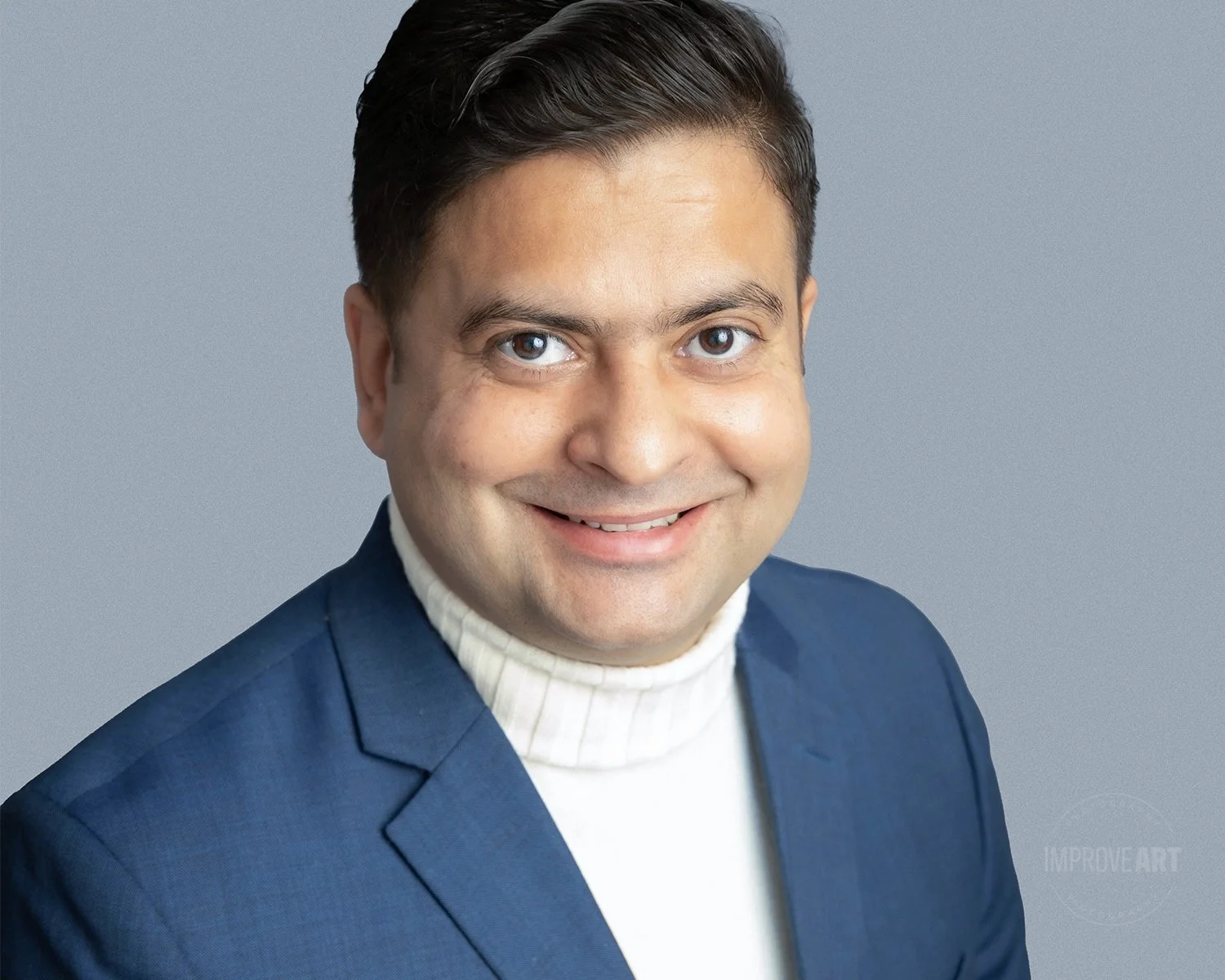 Headshot of a man with dark hair, wearing a navy blazer and white turtleneck sweater, smiling against a gray background.