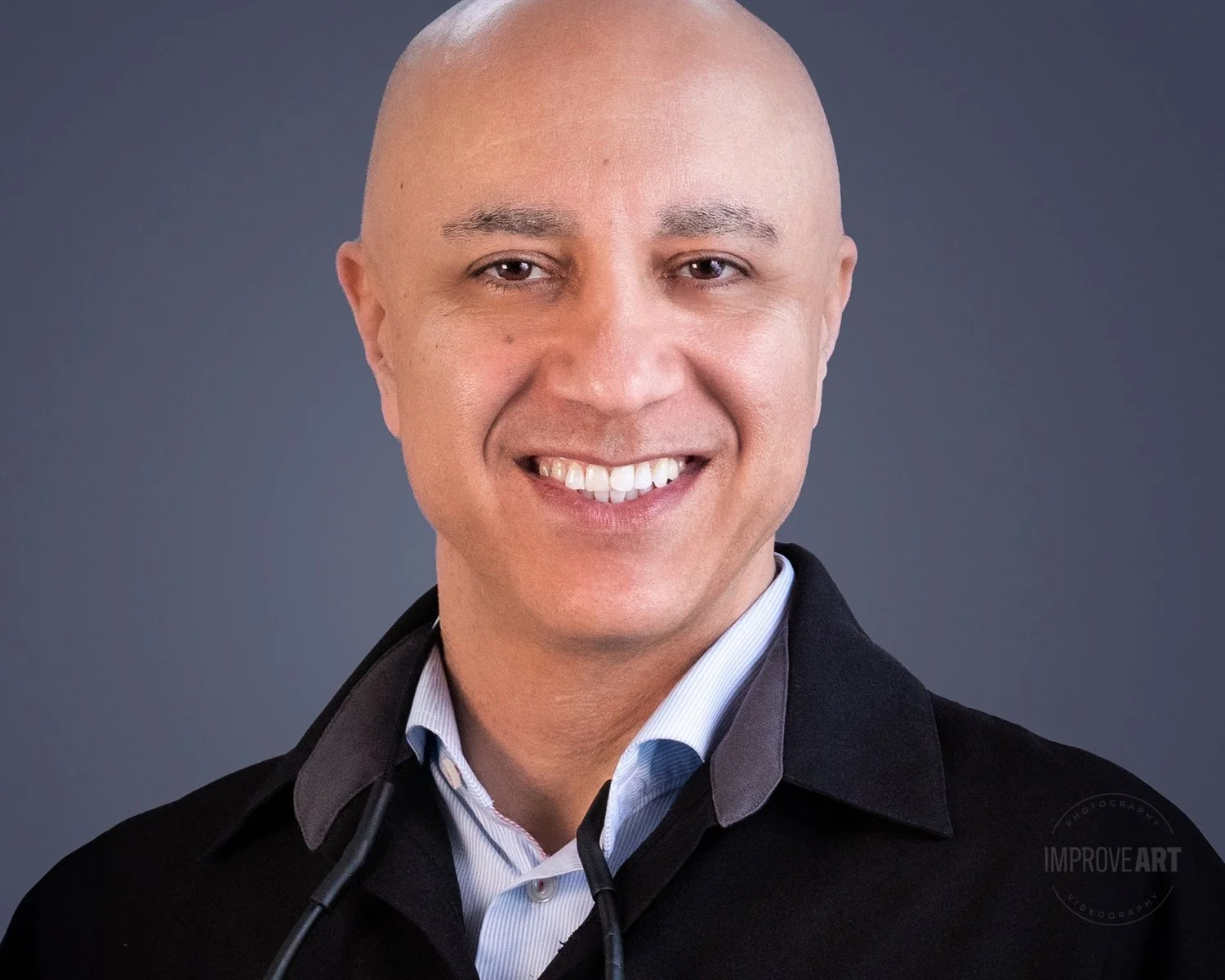 A smiling man with a shaved head wearing a collared shirt and a dark jacket, against a plain gray background.