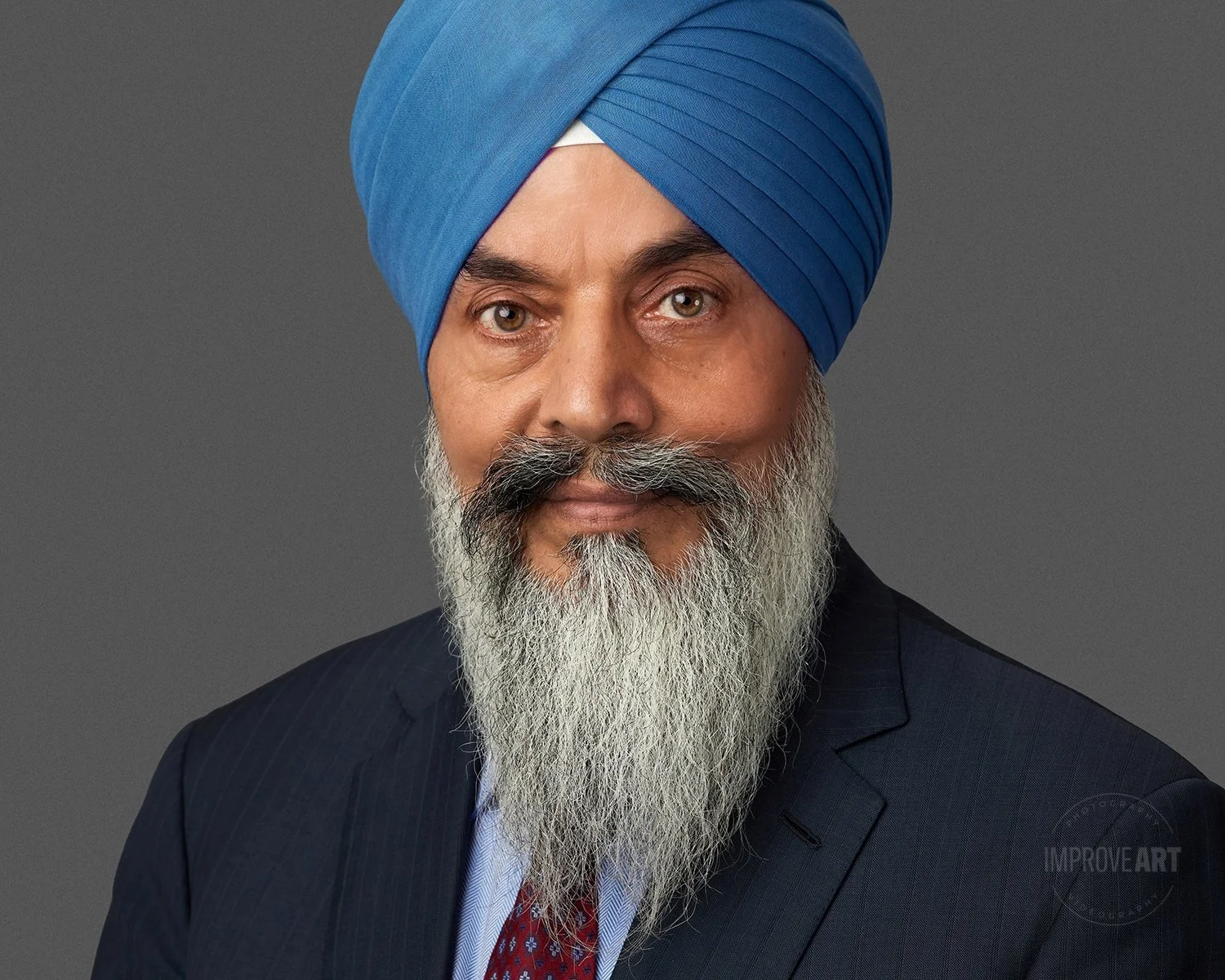 Portrait of a man with a long gray beard wearing a blue turban and a dark suit, looking at the camera against a gray background.