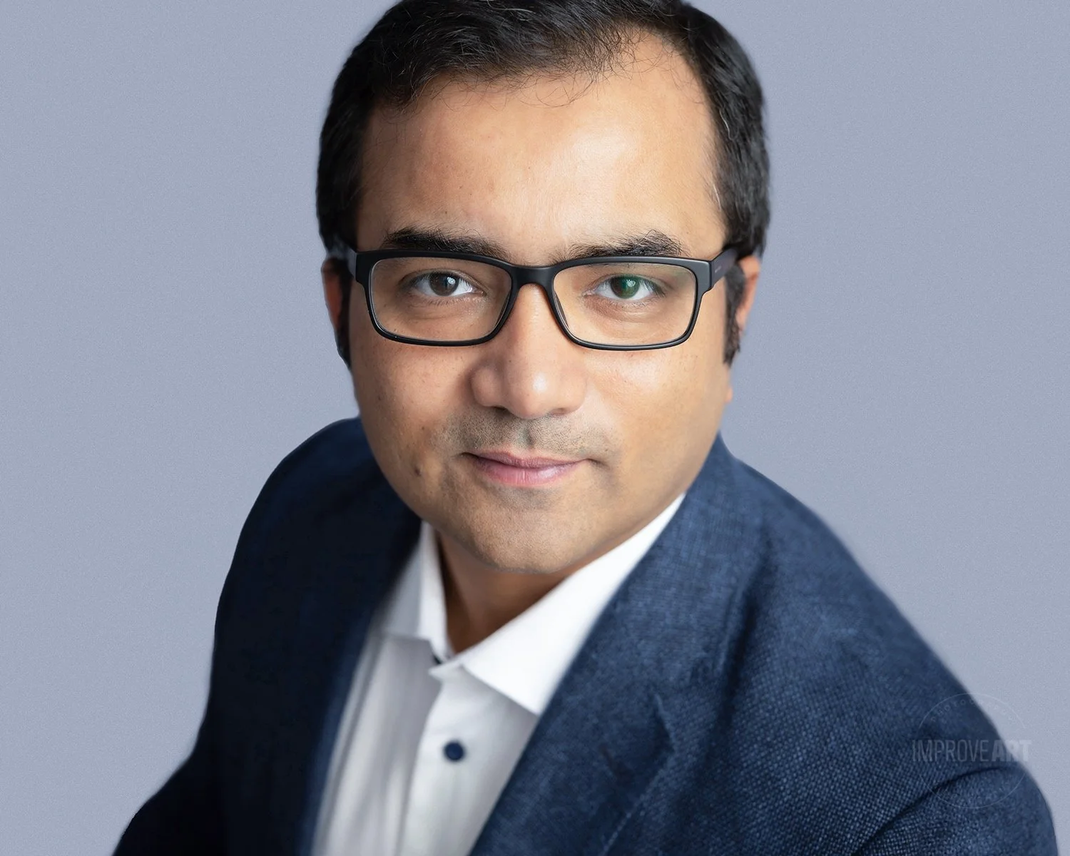A professional headshot of a man wearing glasses, a white shirt, and a blue blazer, looking at the camera with a neutral expression against a plain grey background.