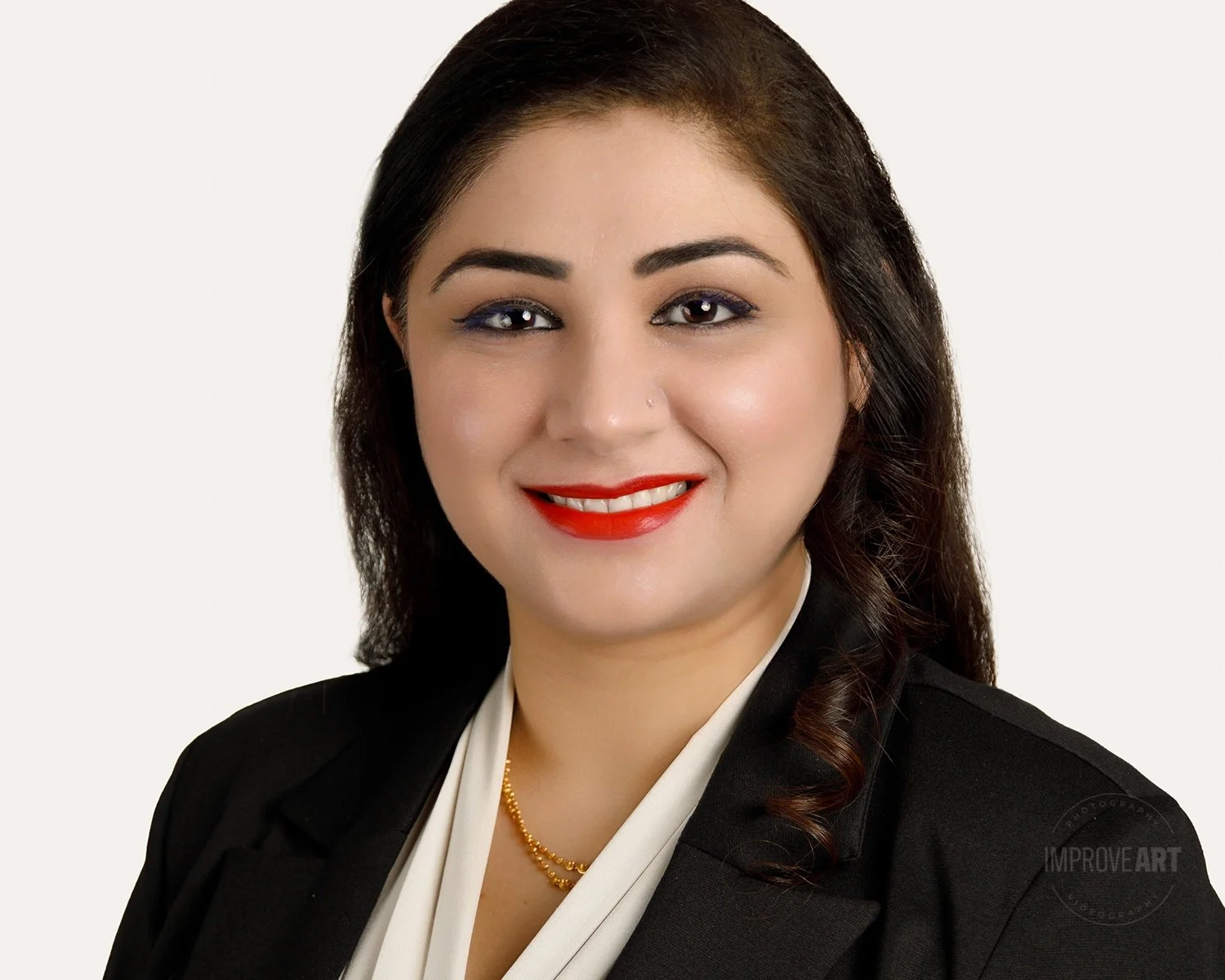 A professional woman with dark brown hair, wearing a black blazer, white blouse, gold necklace, and red lipstick, smiling against a plain white background.
