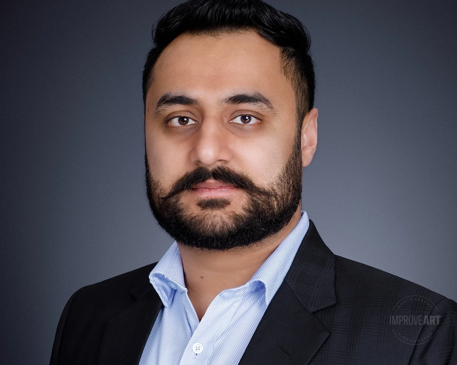 Portrait of a man with dark hair and a beard, wearing a light blue dress shirt and a dark blazer, against a dark background.