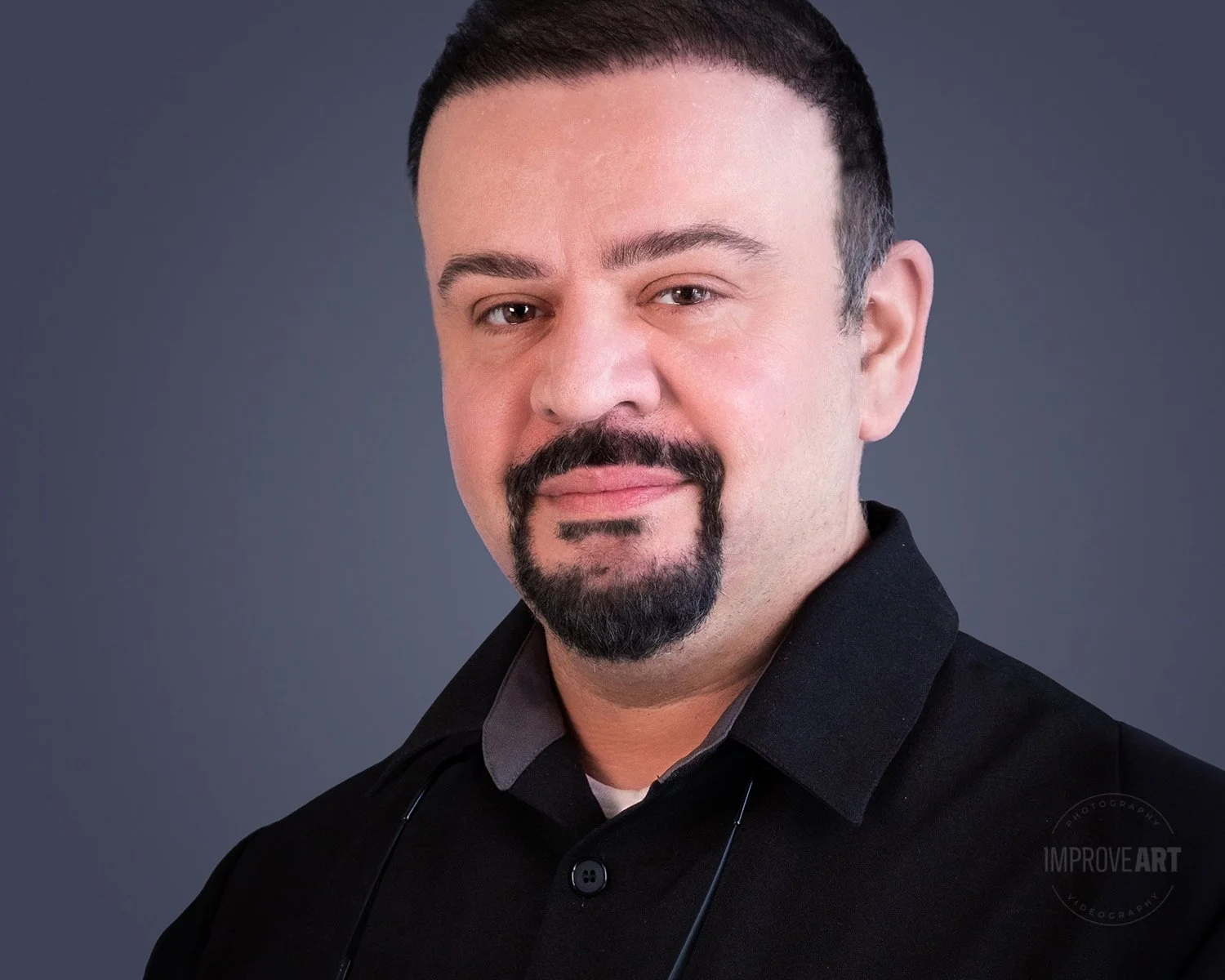 A man with short dark hair, a mustache and goatee, wearing a black collared shirt, posed against a gray background.