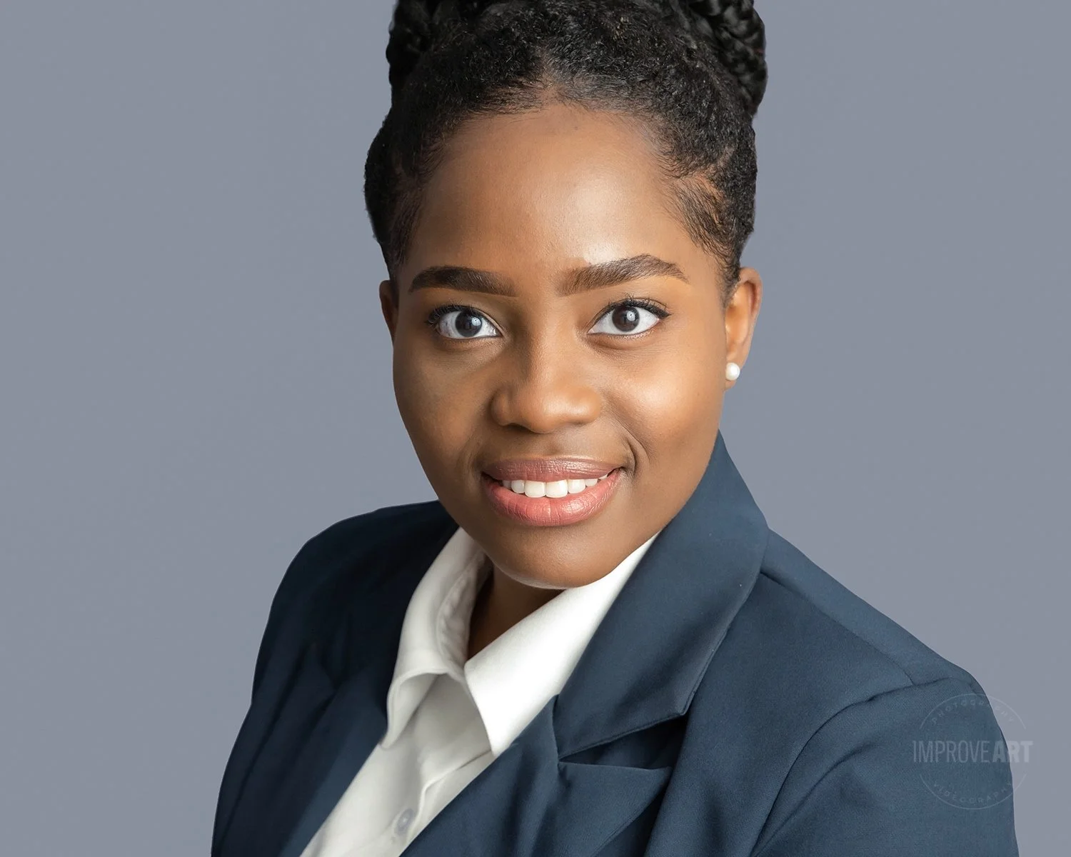 Professional woman with dark braided hair, wearing a dark blazer and white shirt, smiling against a gray background.