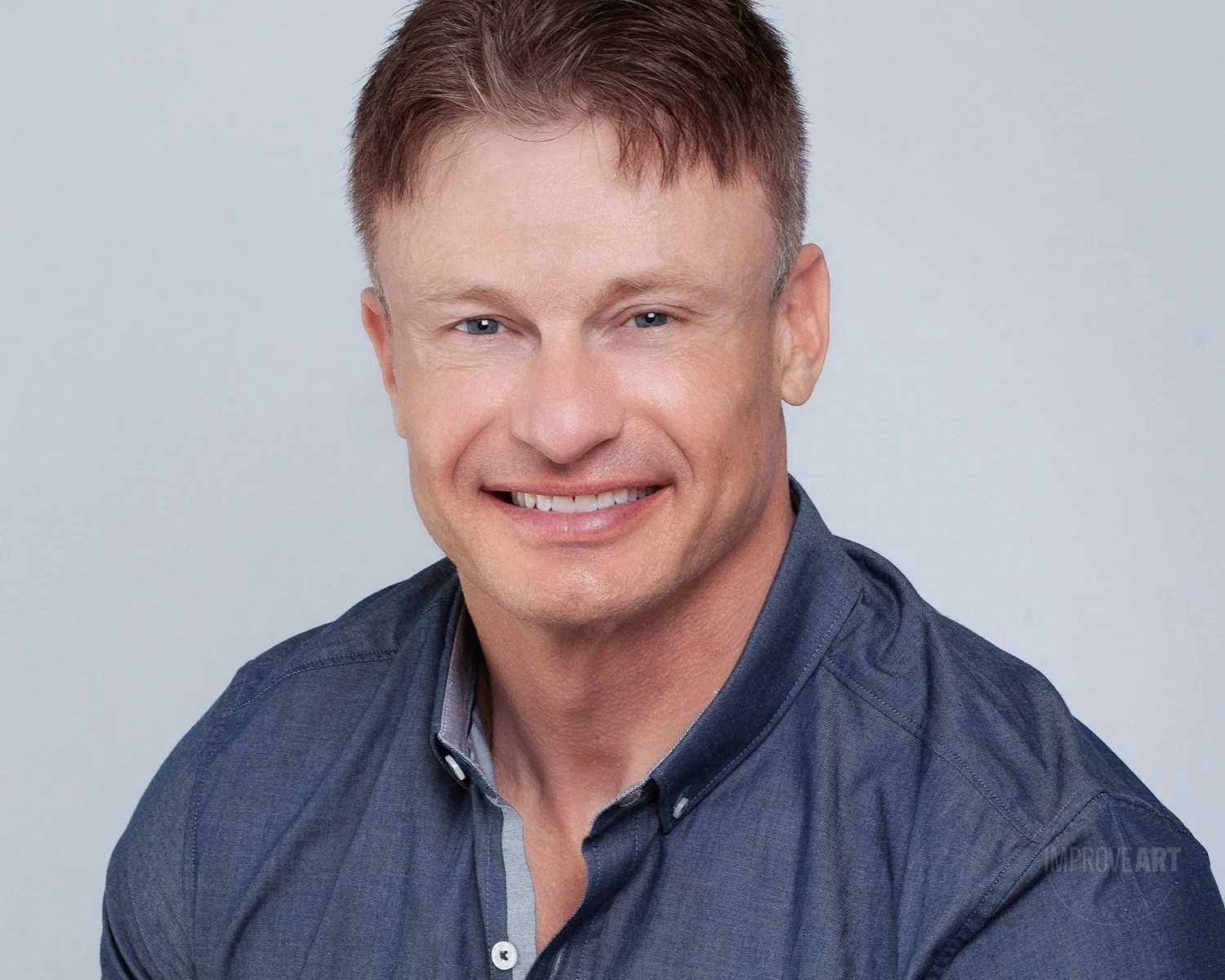 Headshot of a smiling man with short light brown hair, wearing a dark blue shirt, against a plain light background.
