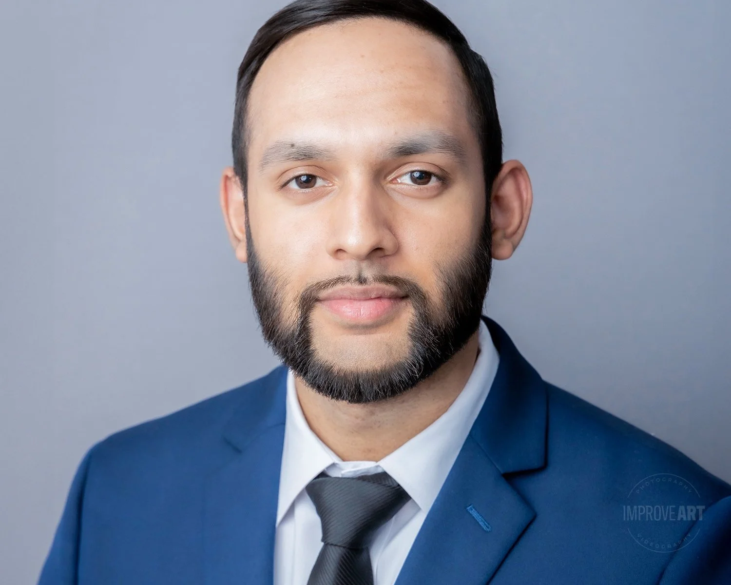 A professional headshot of a man with dark hair and a beard, wearing a blue suit, white shirt, and black tie, against a plain gray background.