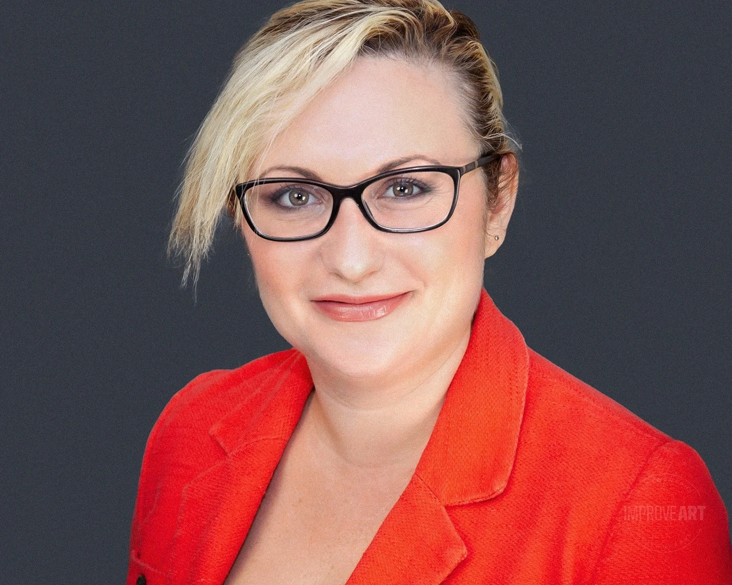 A woman with short blonde hair, glasses, and light makeup, wearing a red blazer, smiling at the camera against a dark grey background.