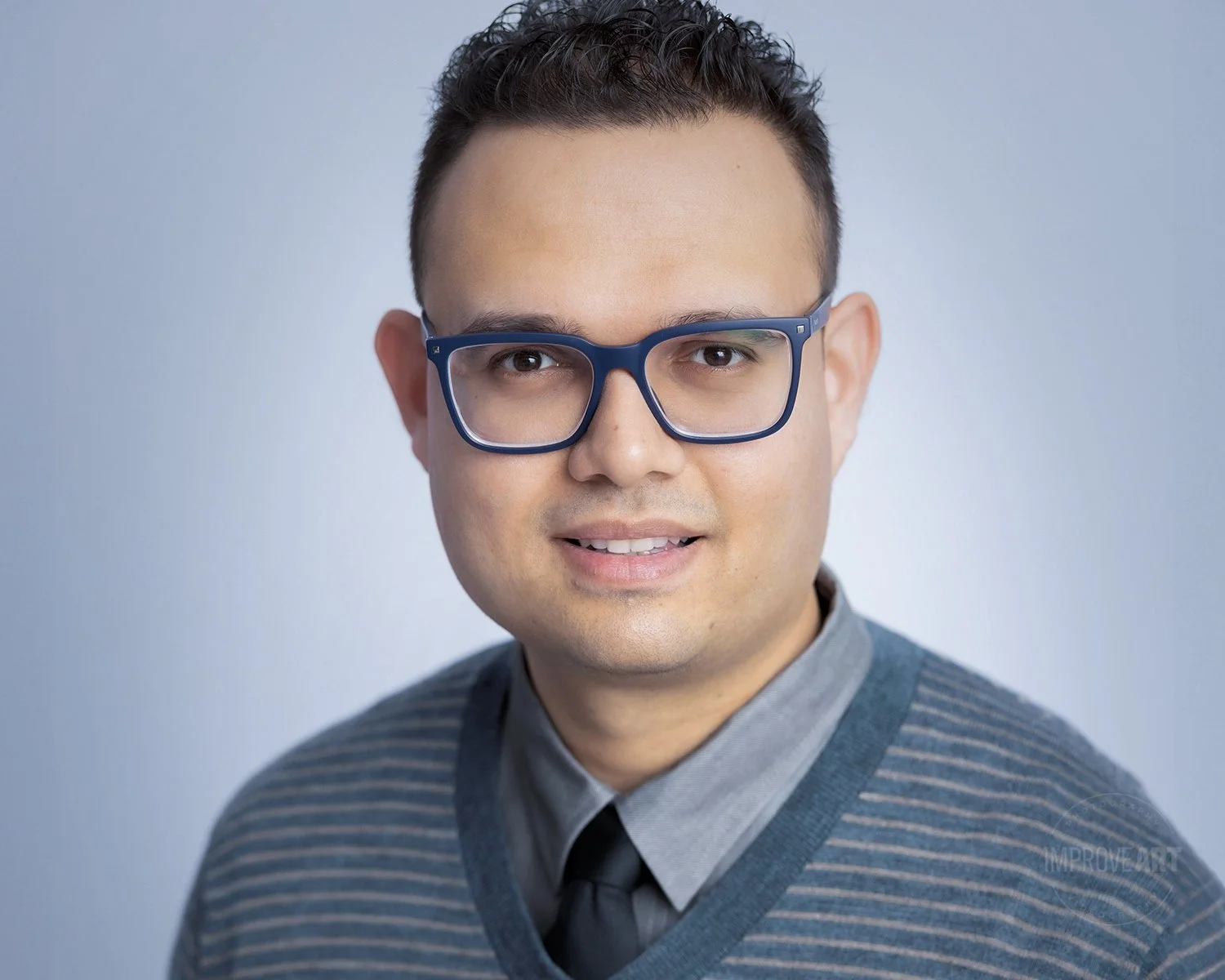 A young man with short dark hair, wearing large blue glasses, a grey collared shirt, a dark grey tie, and a striped sweater, smiling at the camera against a light grey background.