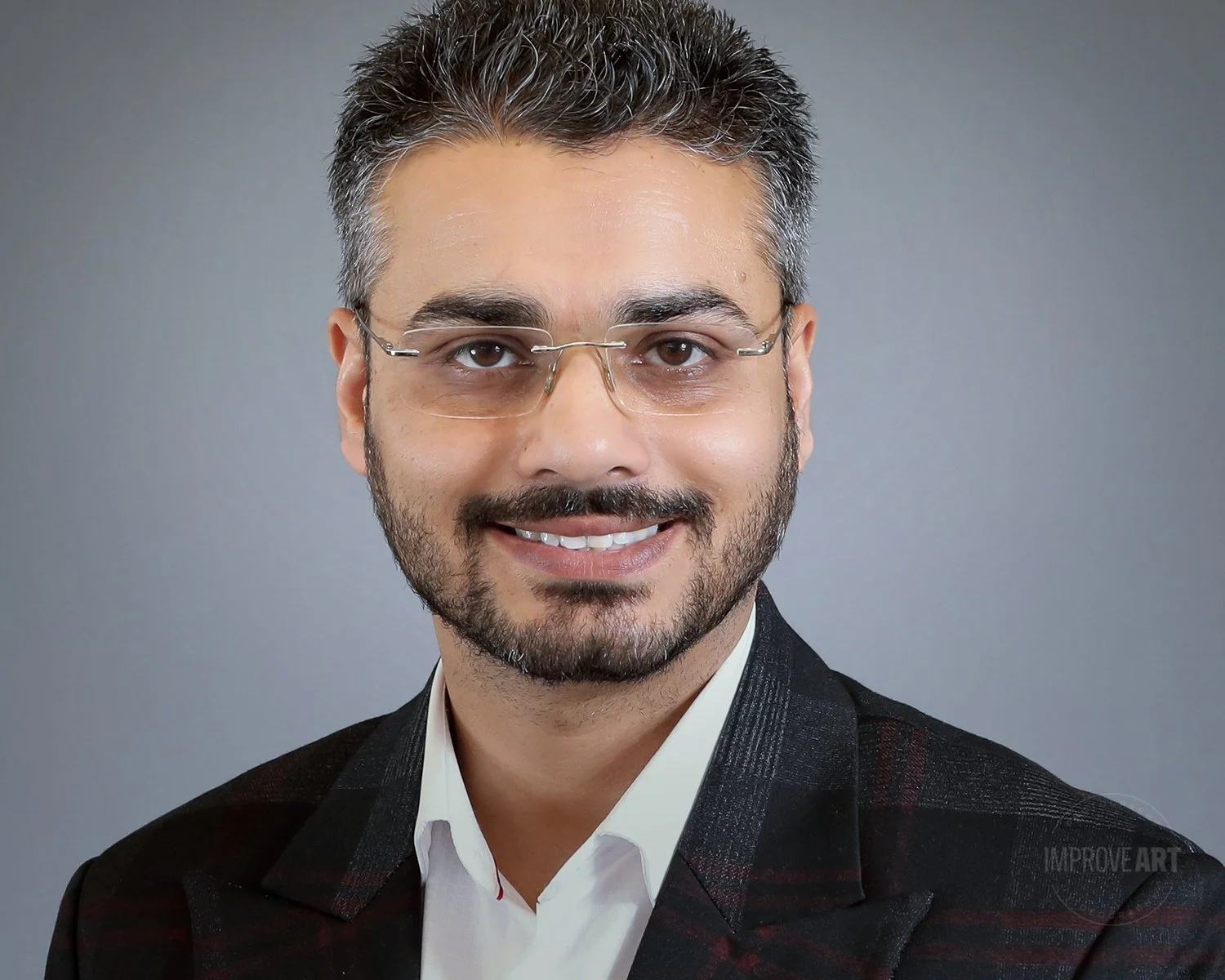 A middle-aged man with dark hair and glasses smiling, wearing a black blazer and white shirt, against a gray background.