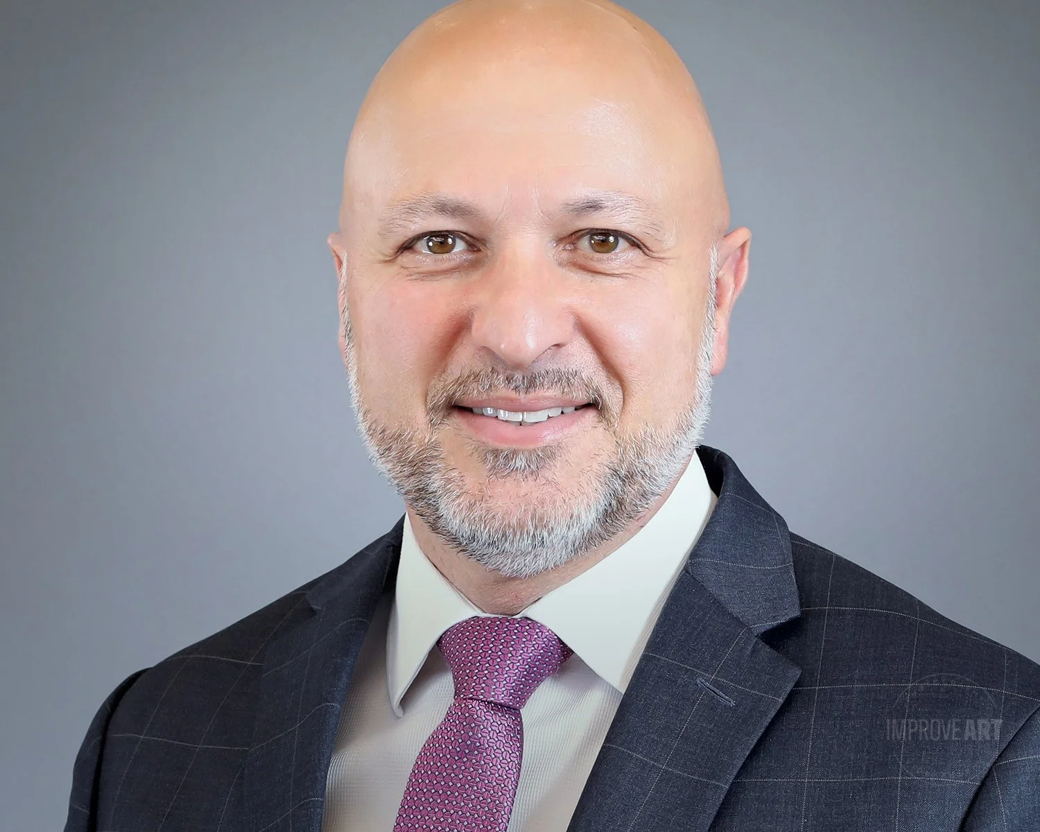 Headshot of a smiling bald man with a beard wearing a suit and tie, against a gray background.