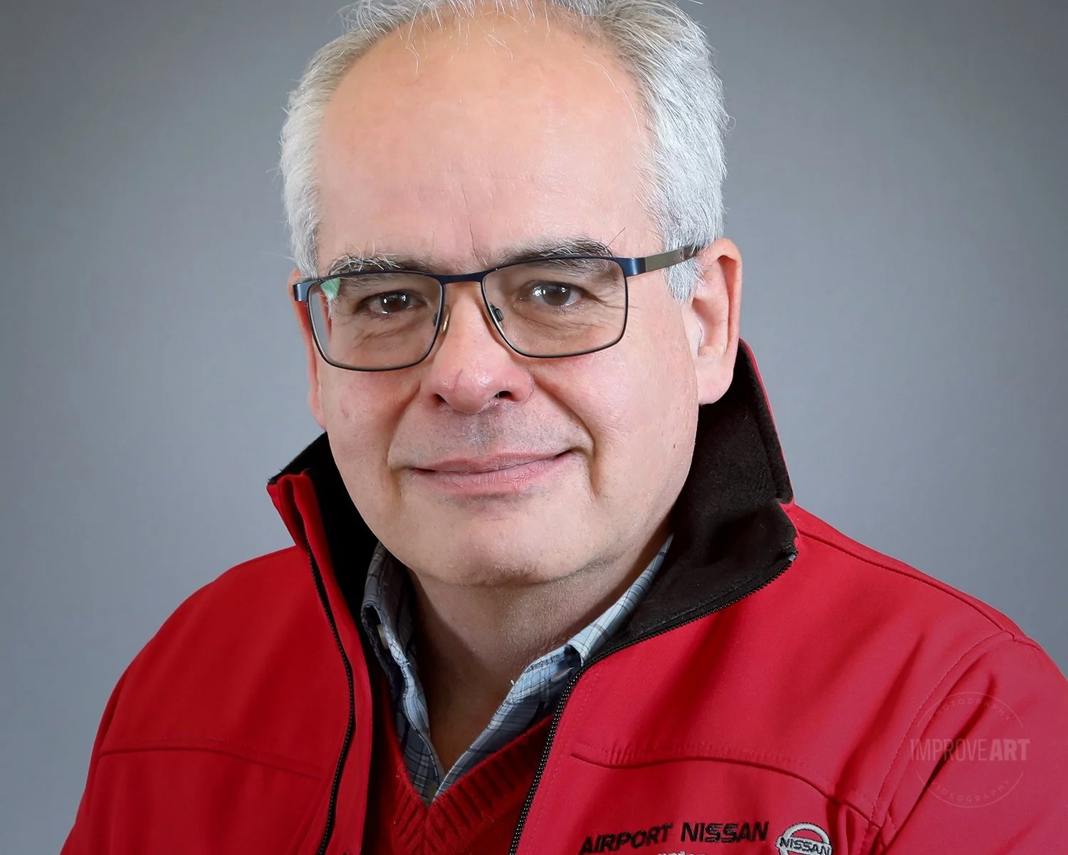 A middle-aged man with gray hair and glasses wearing a red jacket with the logo 'Airport Nissan'.