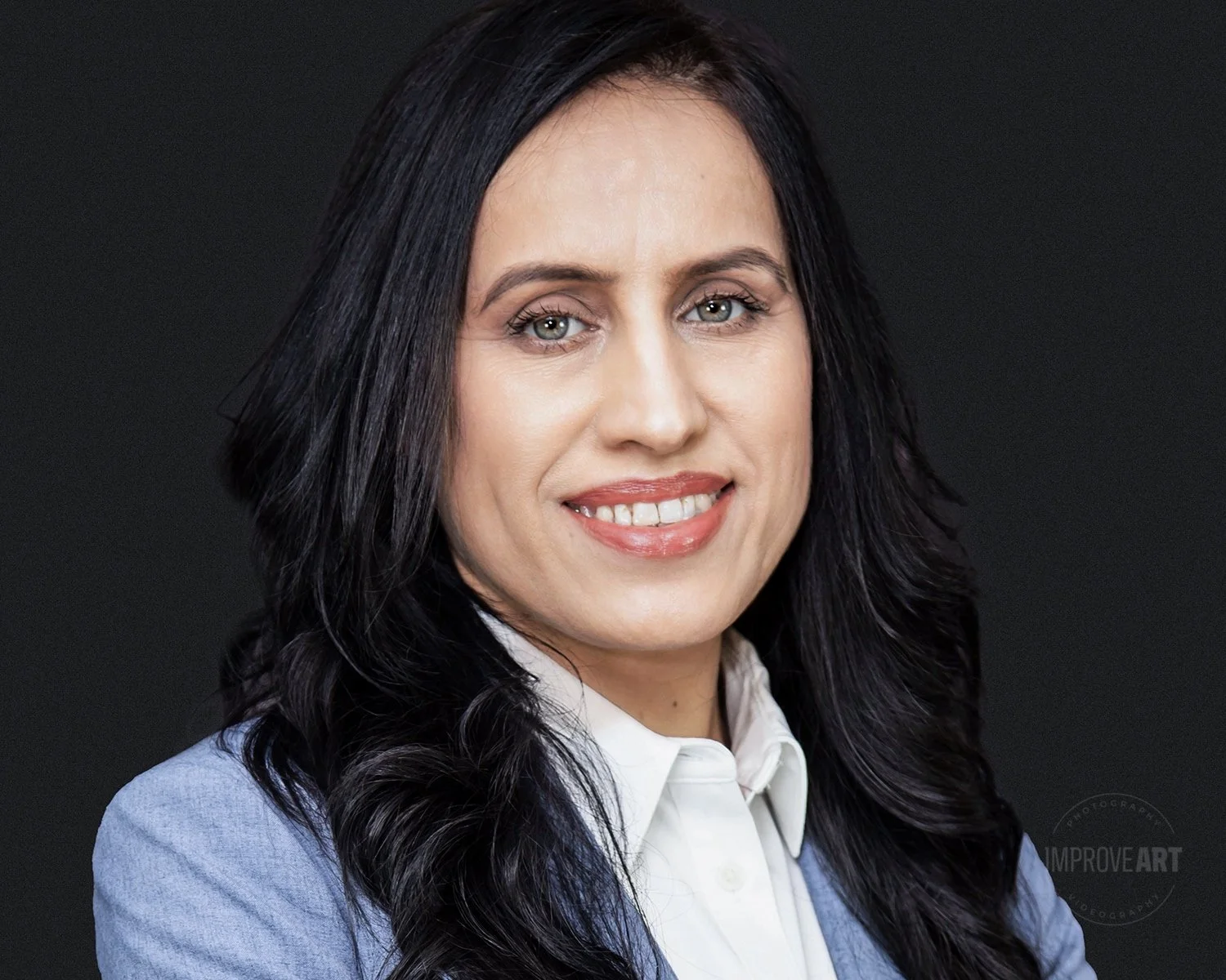 Professional woman with long dark wavy hair, wearing a white shirt and blue blazer, smiling against a dark grey background.