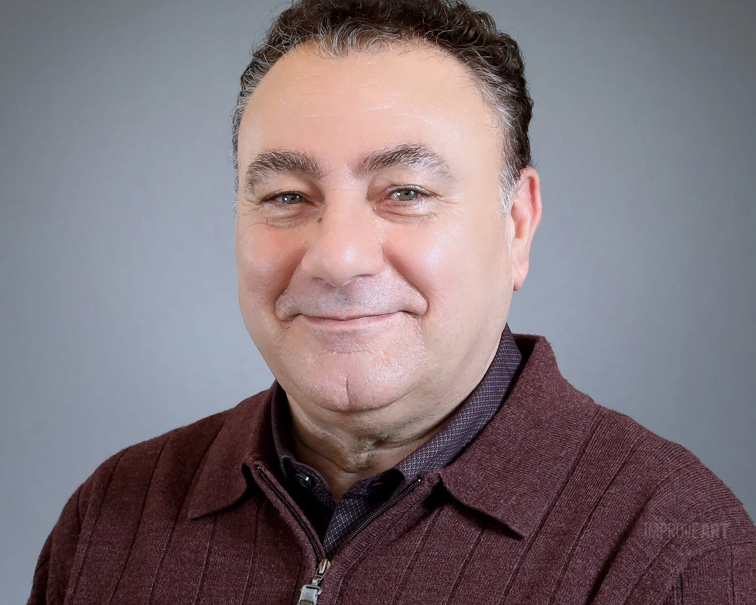 A middle-aged man with short, curly dark hair and light eyes, smiling softly, wearing a maroon zip-up sweater and a dark collared shirt underneath, standing against a plain gray background.