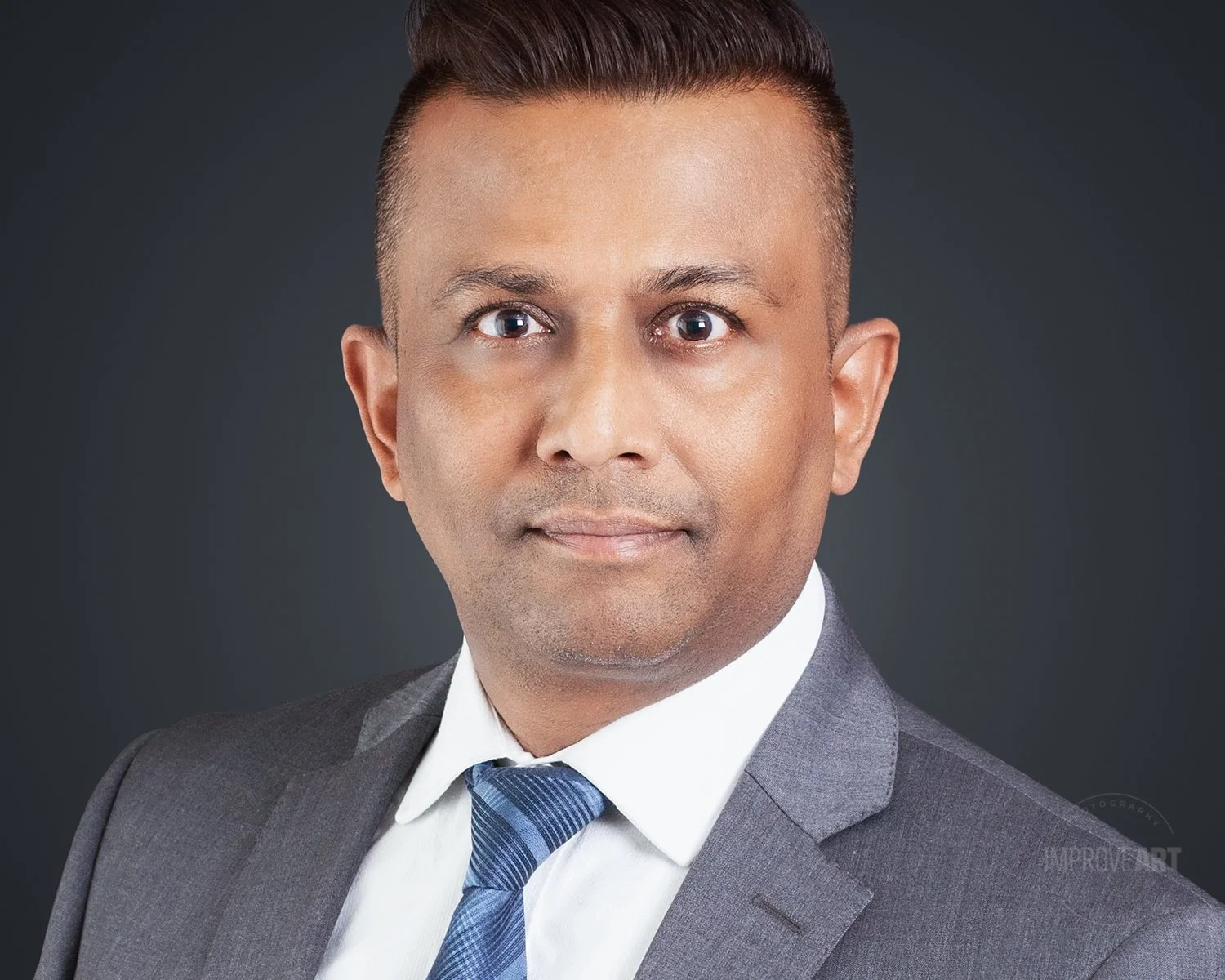 Professional portrait of a man in a gray suit, white shirt, and blue tie, against a dark background.