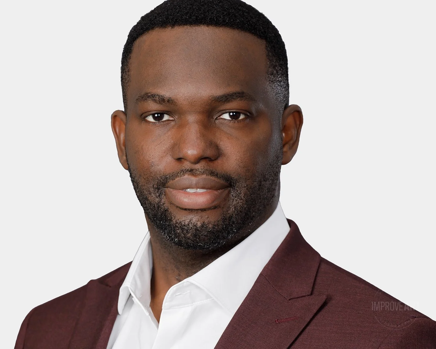 Close-up portrait of an African American man with short hair and a beard, wearing a white shirt and maroon blazer against a plain light background.