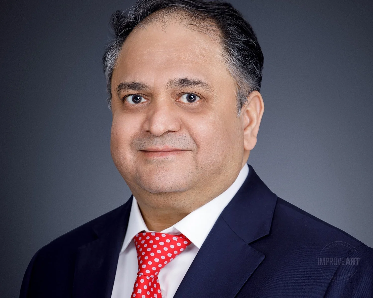 A portrait of a middle-aged man with dark hair streaked with gray, wearing a navy suit, white shirt, and a red polka dot tie, against a gray background.