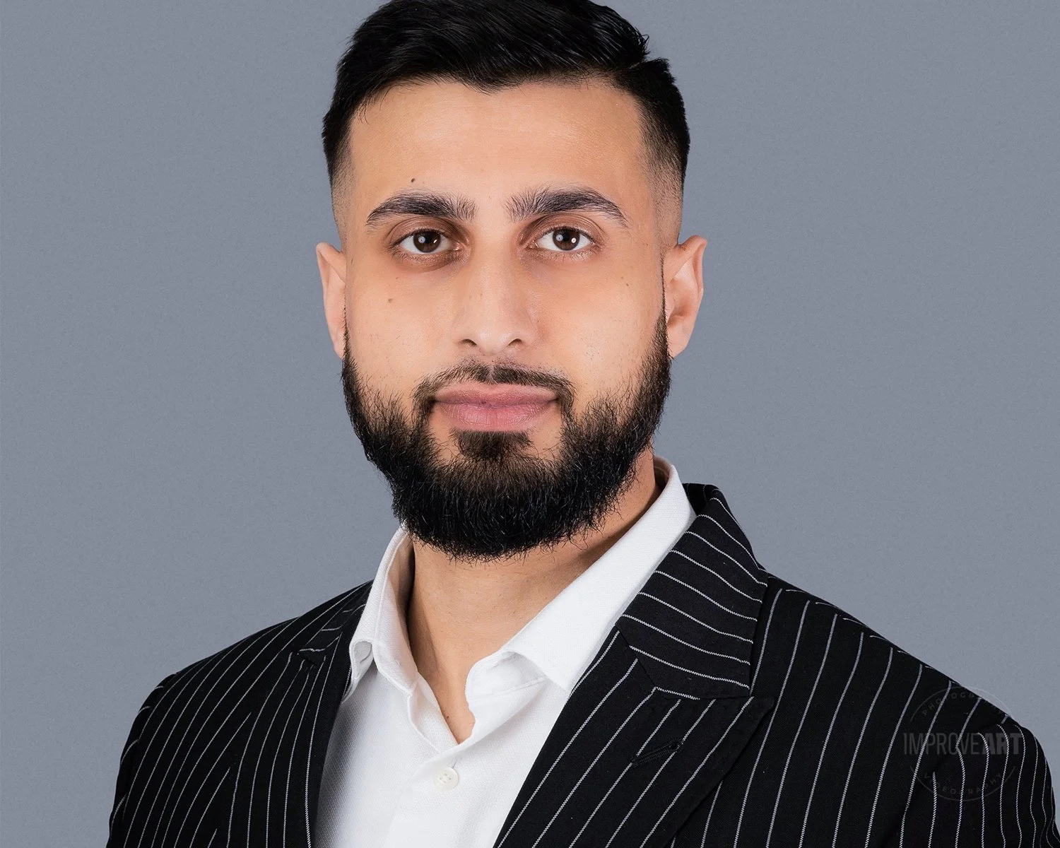 A portrait of a man with short dark hair and a full beard, wearing a white shirt and a black pinstripe suit, against a plain gray background.