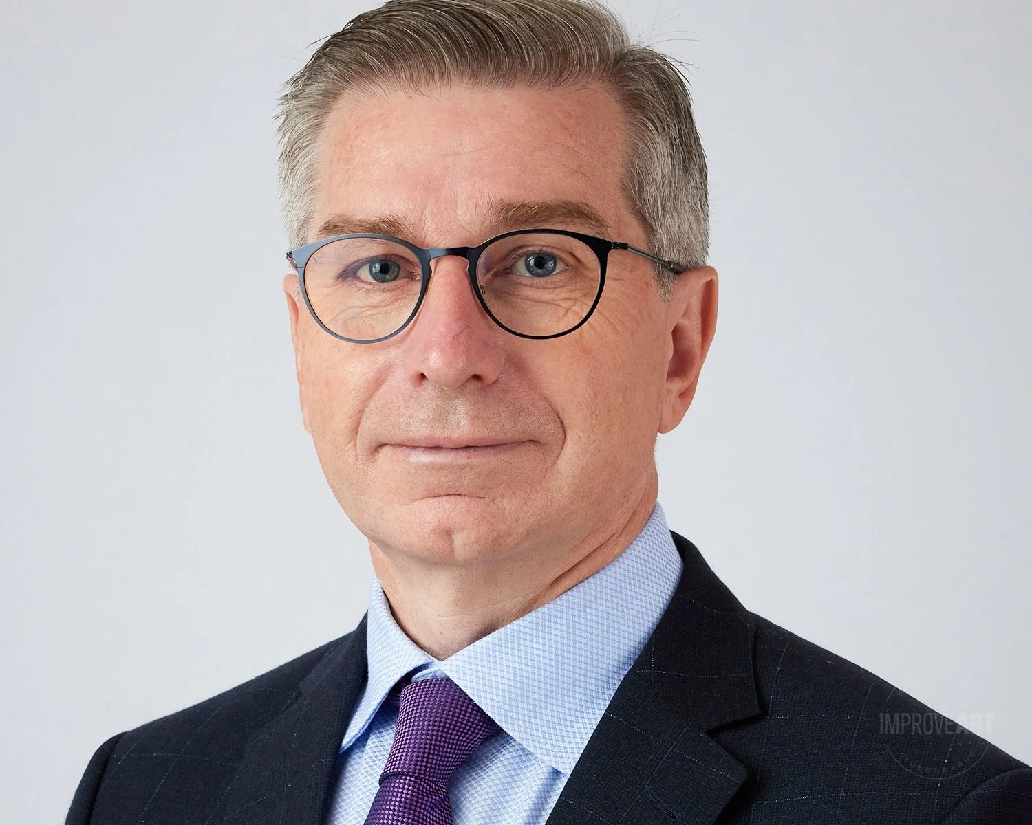 Professional portrait of a middle-aged man with glasses, wearing a dark suit, light blue shirt, and purple tie, against a plain light background.