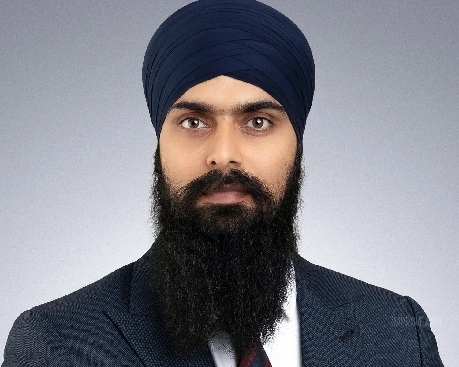 A man wearing a dark blue turban, a dark suit, and a white shirt with a maroon tie, sporting a thick black beard, against a light gray background.