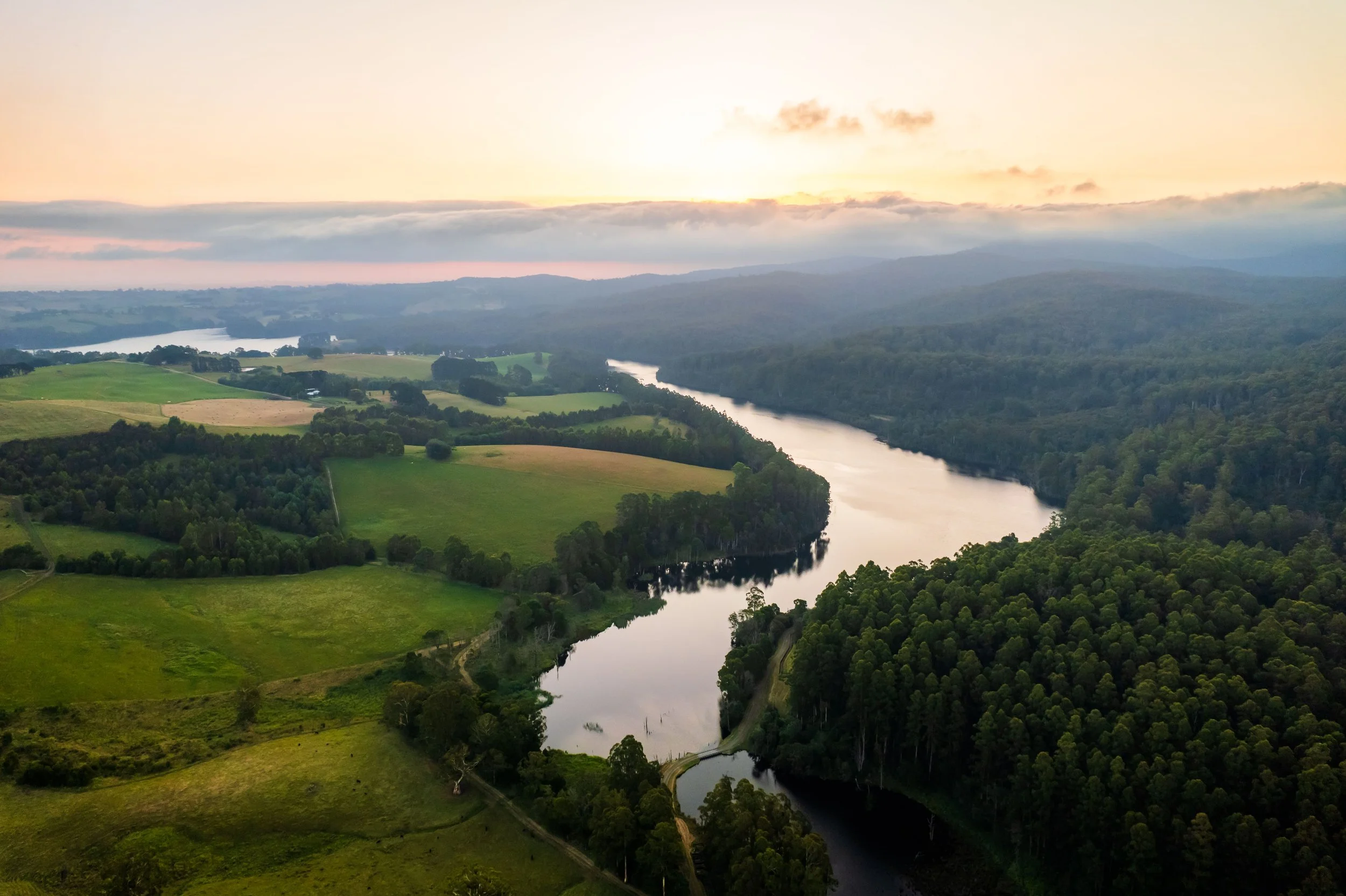 A river winding through green countryside with rolling hills and forested areas, under a sky with scattered clouds at sunset or sunrise.
