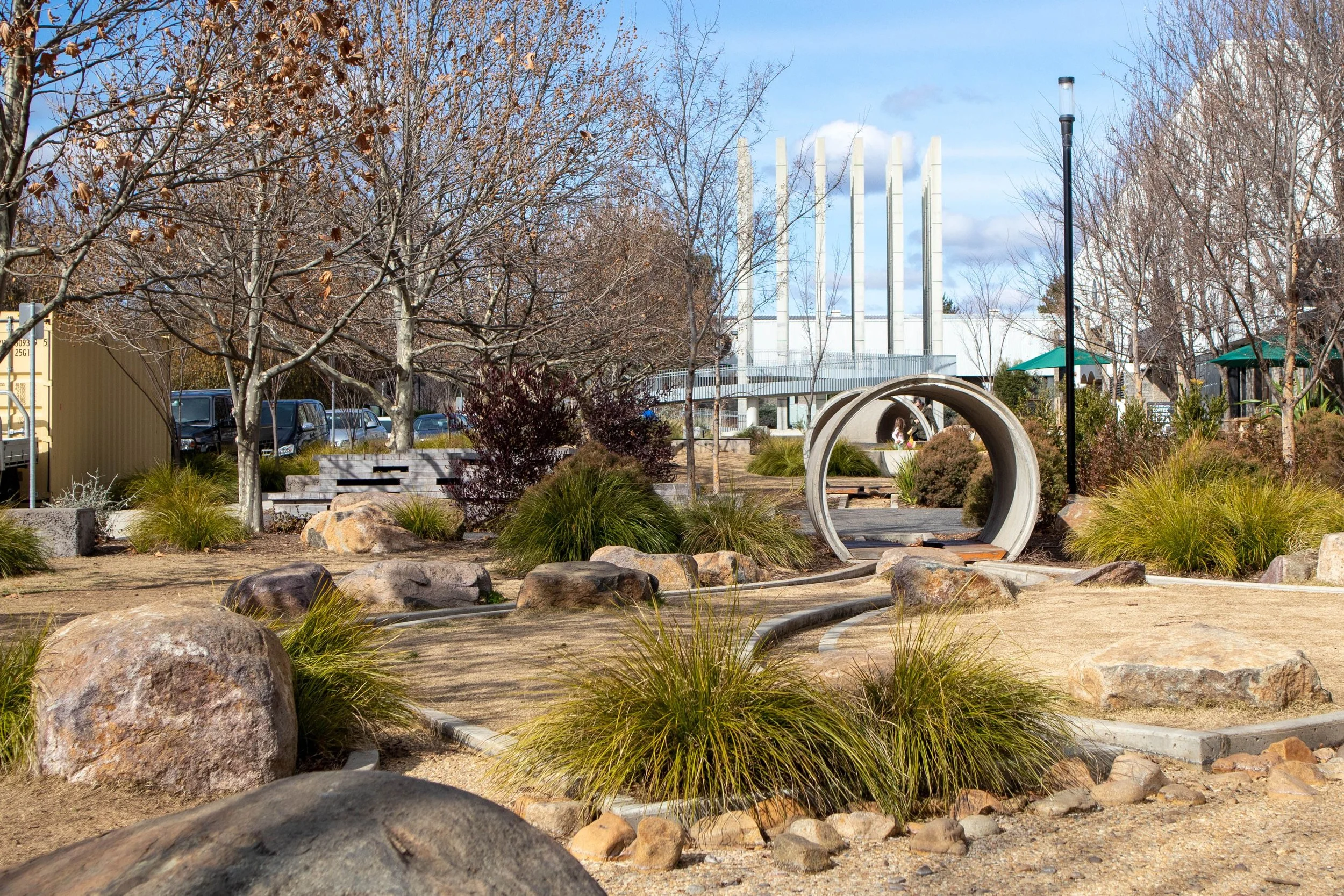 An outdoor park area with rocks, native plant, native trees, native grasses and a concrete tunnel, with a modern industrial building and a parking lot in the background.