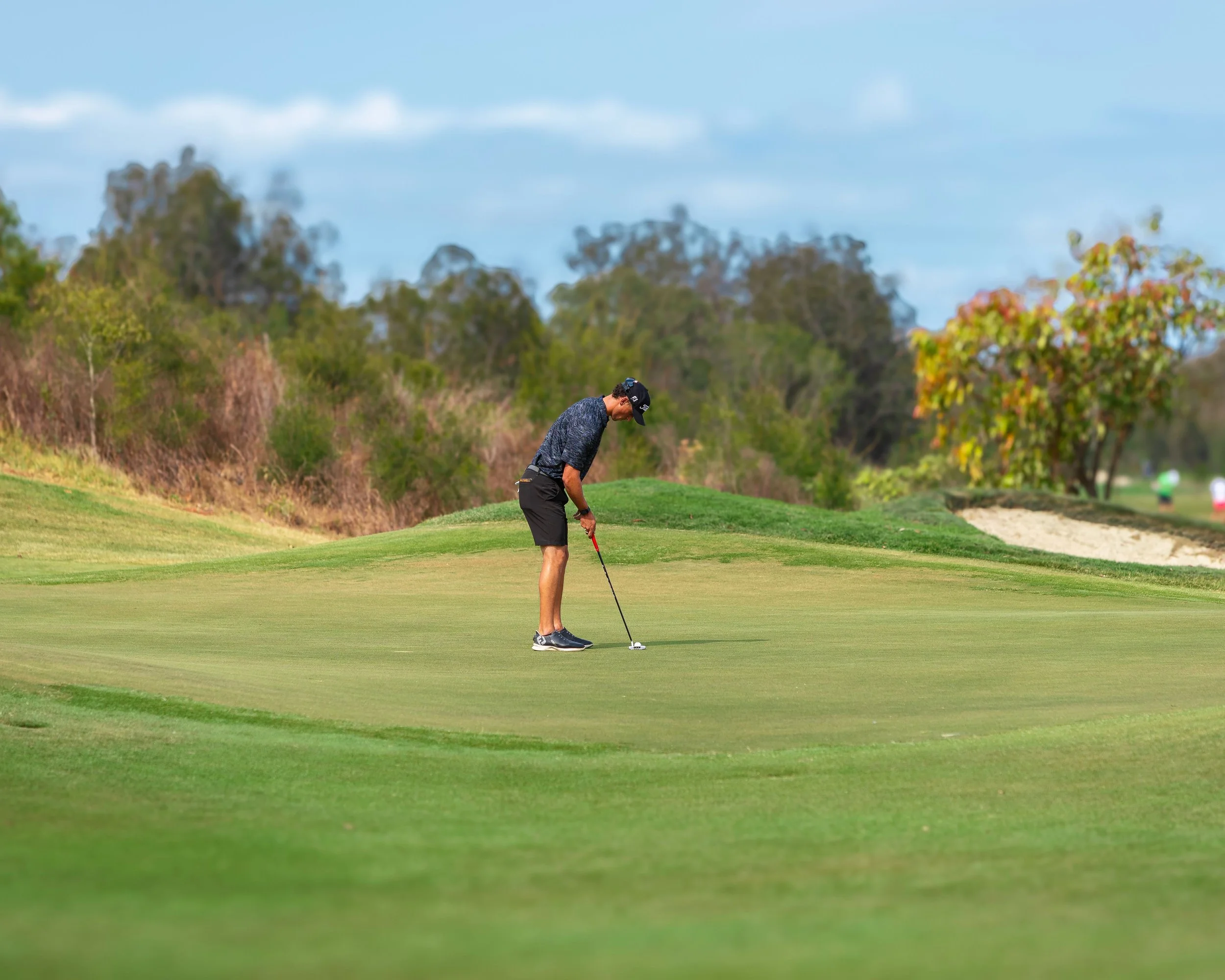A person in a dark shirt and shorts prepares to hit a golf ball on a putting green with trees and blue sky in the background.