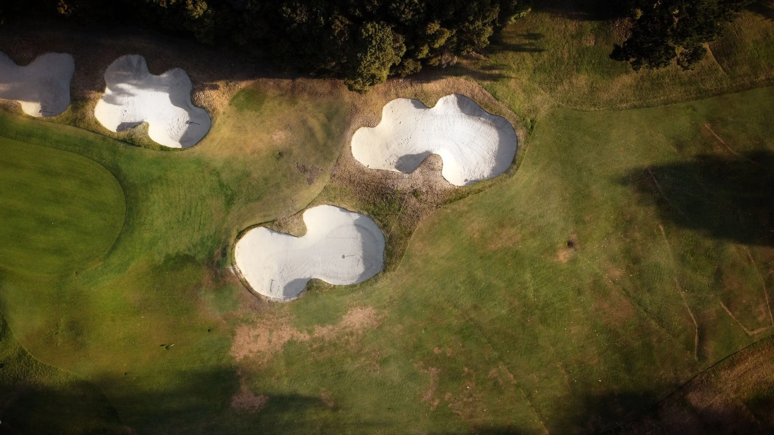An aerial view of a golf course with several sand bunkers and a green surrounded by native trees and native grasses.