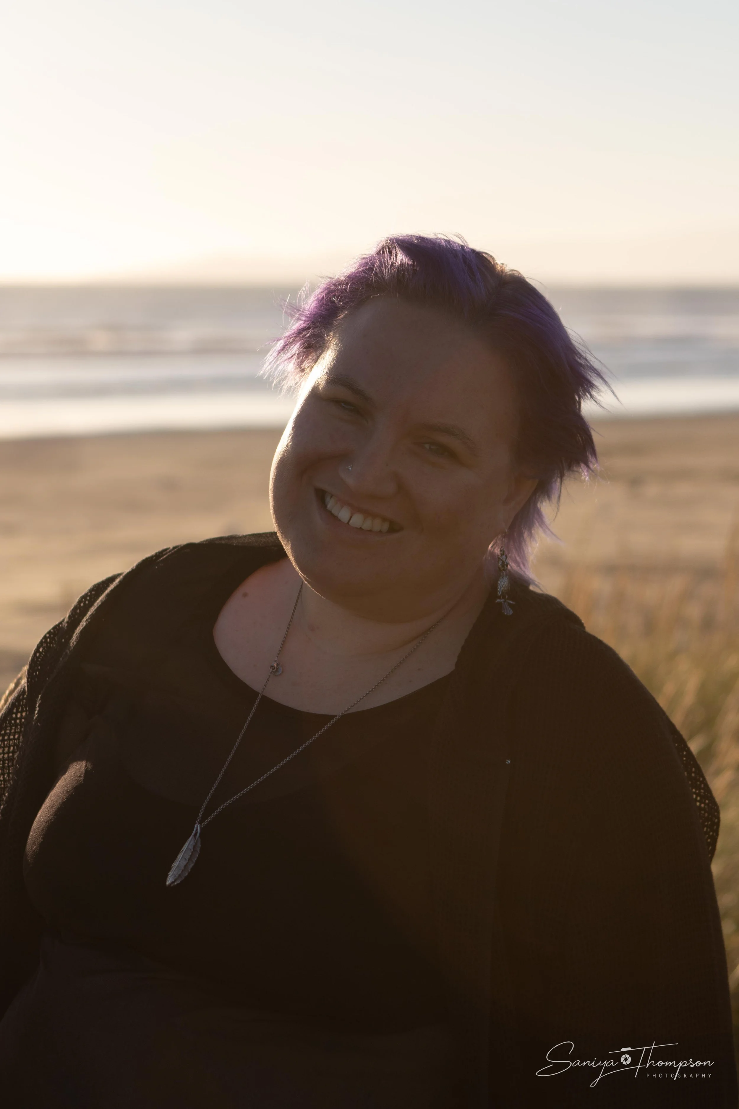 A woman smiling at sunset on a beach with the ocean in the background, wearing a black top and jewelry, with purple hair.