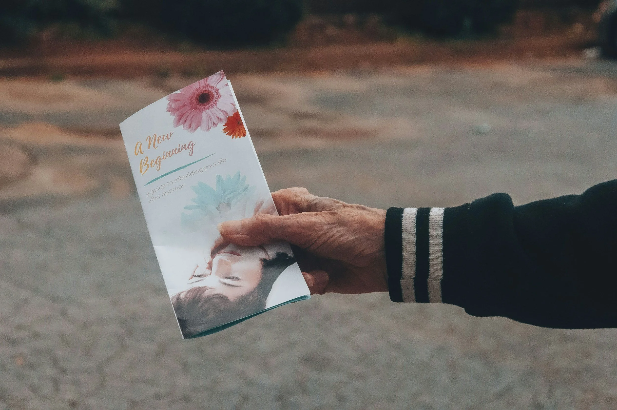 Hand holding a pamphlet titled 'A New Beginning' with floral designs, in an outdoor setting.