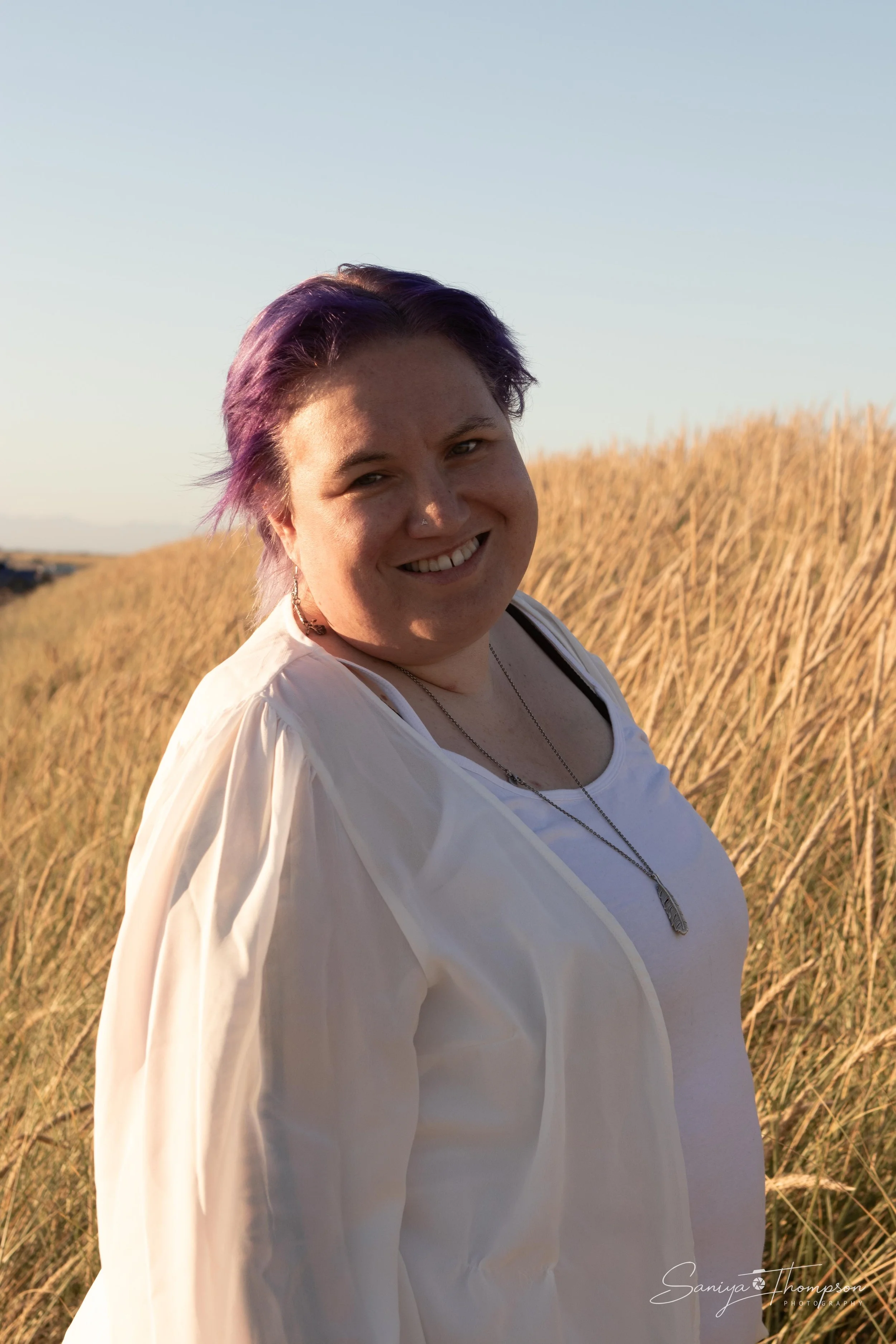 A woman with purple hair smiling outdoors during sunset, wearing a white top and a light-colored jacket, with tall grass in the background.