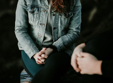 Two people sitting facing each other, one wearing a denim jacket, holding hands.