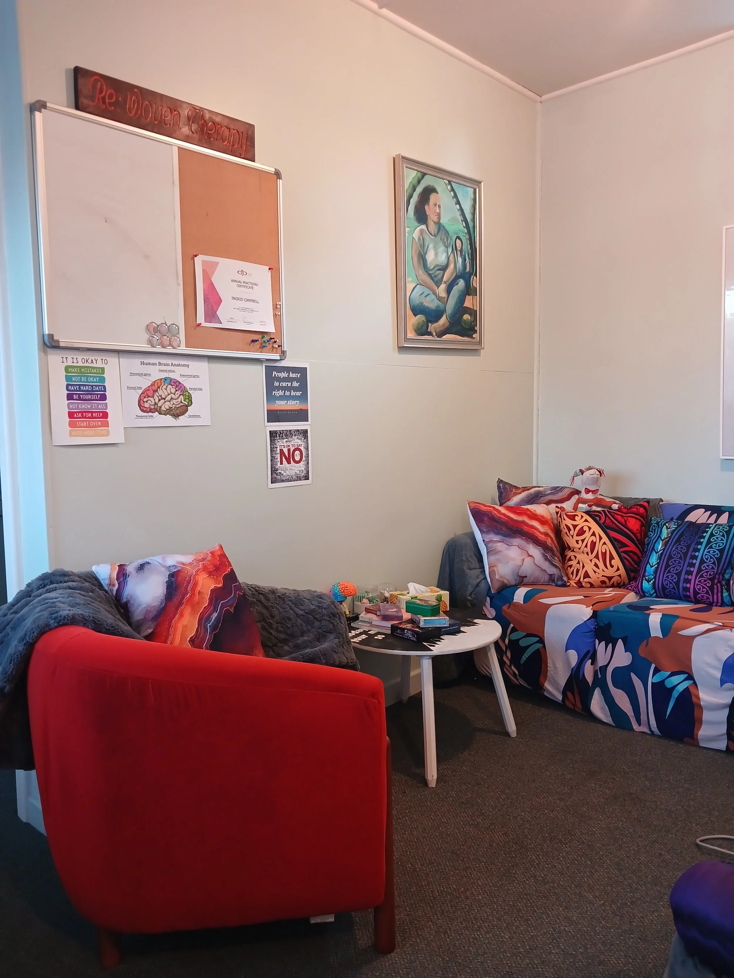 Living room with a red armchair, a sofa with patterned pillows, and a white coffee table. The walls have posters, a painting, and a whiteboard with notes. There are blankets and various items on the table.