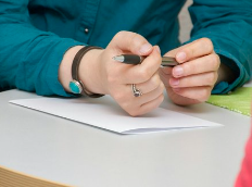 Person writing on a piece of paper with a pen, wearing rings and a teal shirt