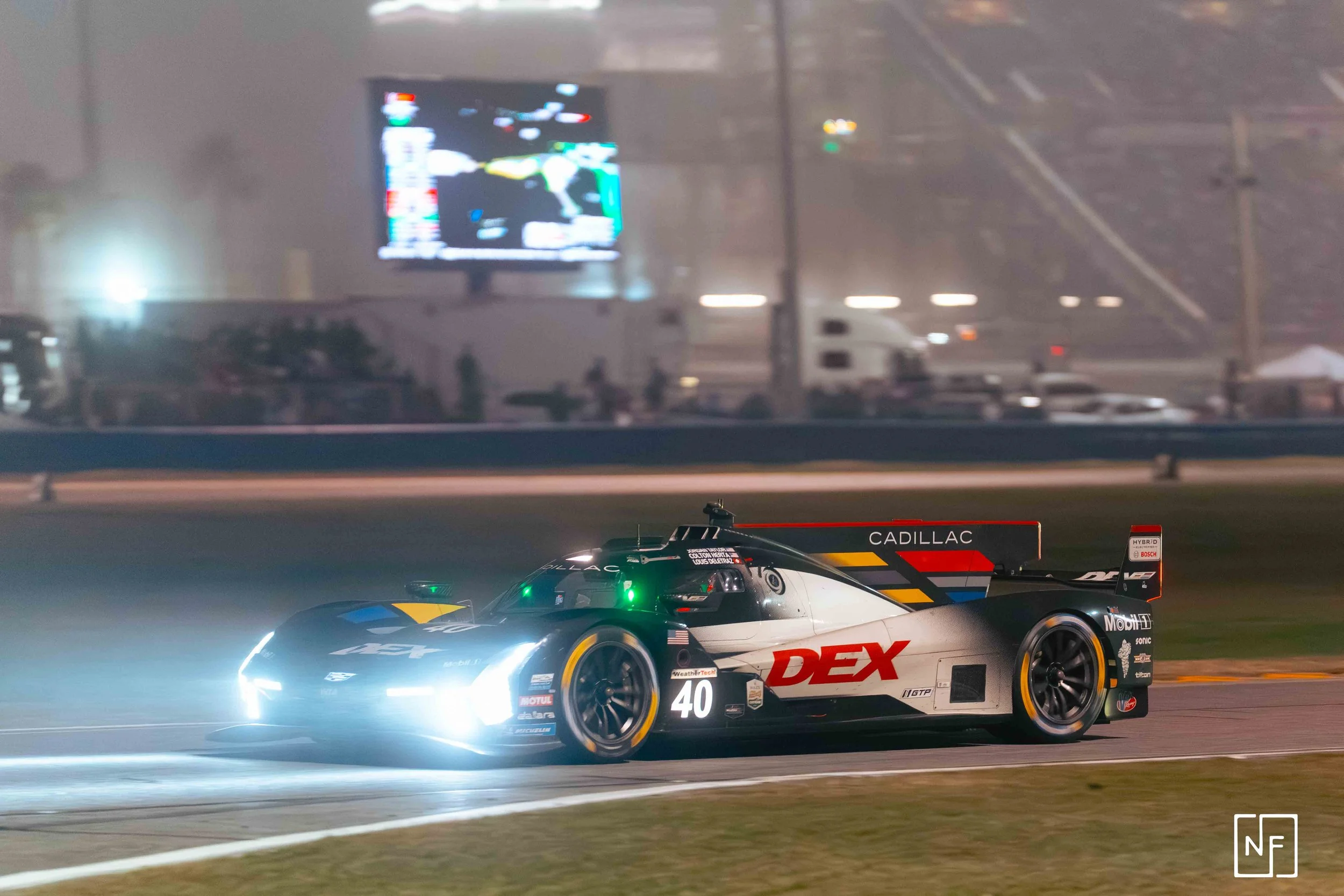A race car on a track at night, headlights on, with a digital billboard in the background.