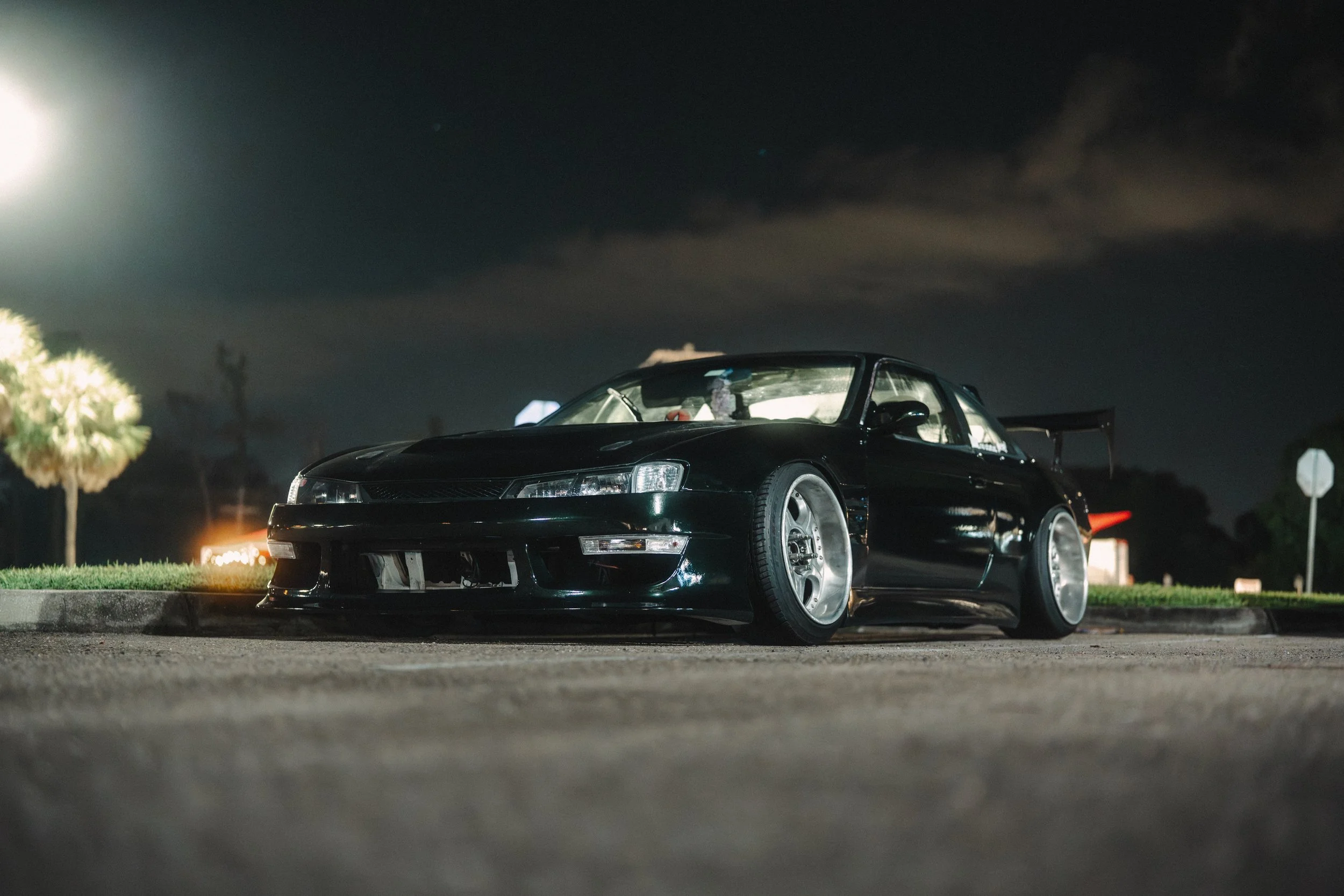 A black sports car parked on the side of a street at night under dark cloudy skies, with a dark background and some trees visible in the distance.