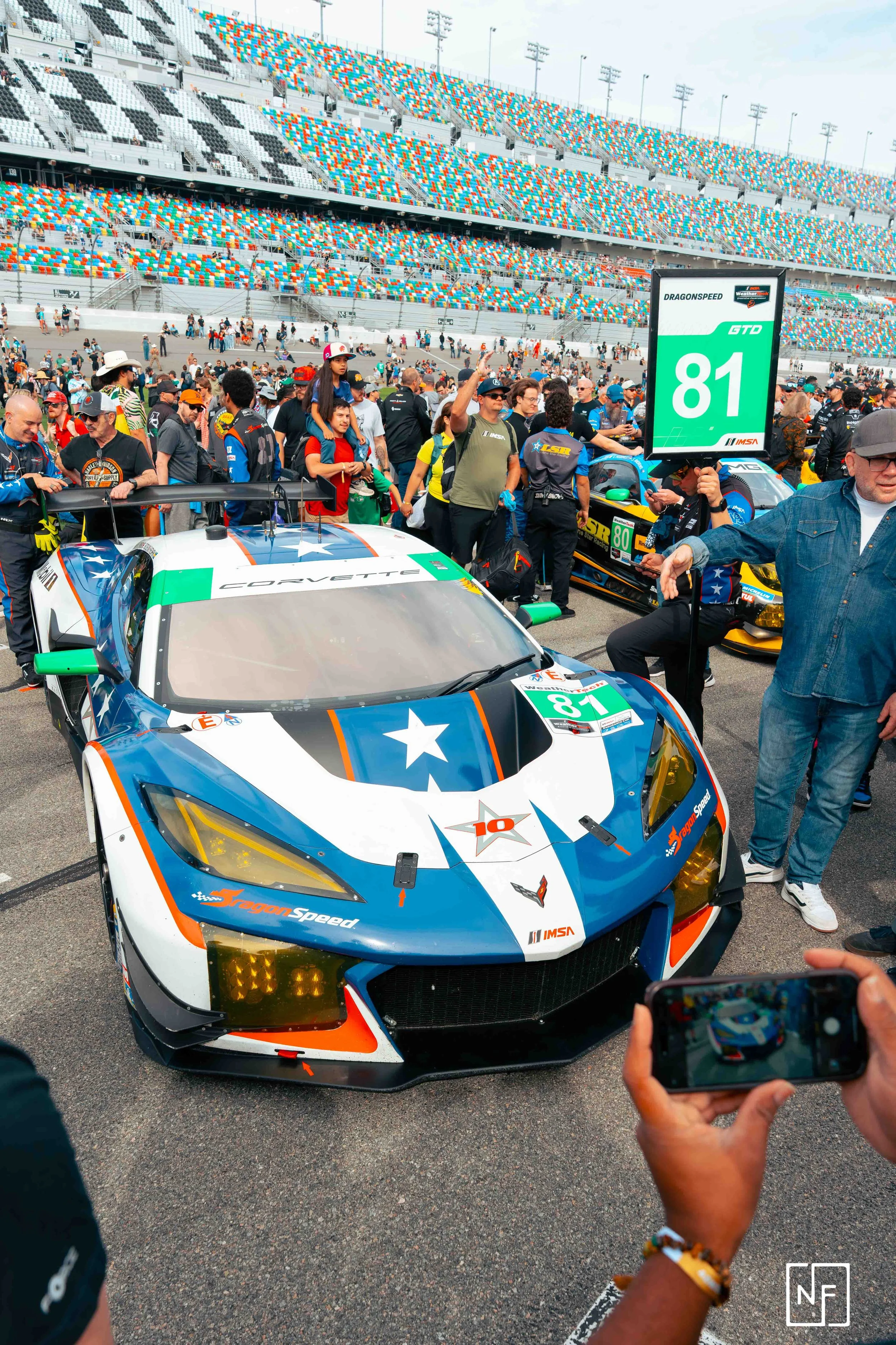 A race car with blue, white, and orange coloring on display at a motorsport event, surrounded by spectators and race officials, with a digital leaderboard showing the number 81.
