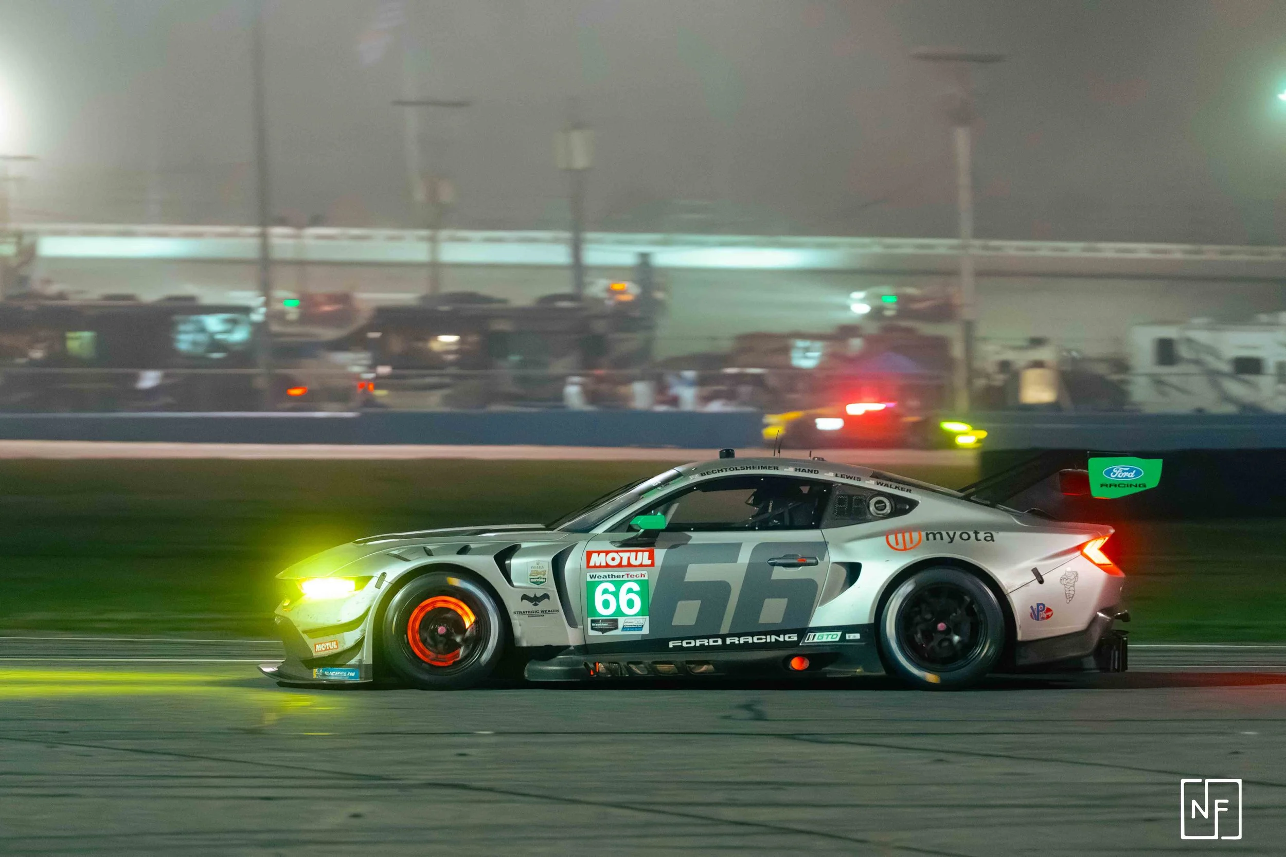 Race car speeding on track at night with a blurred background of vehicles and spectators, marked with sponsors and race number 66.