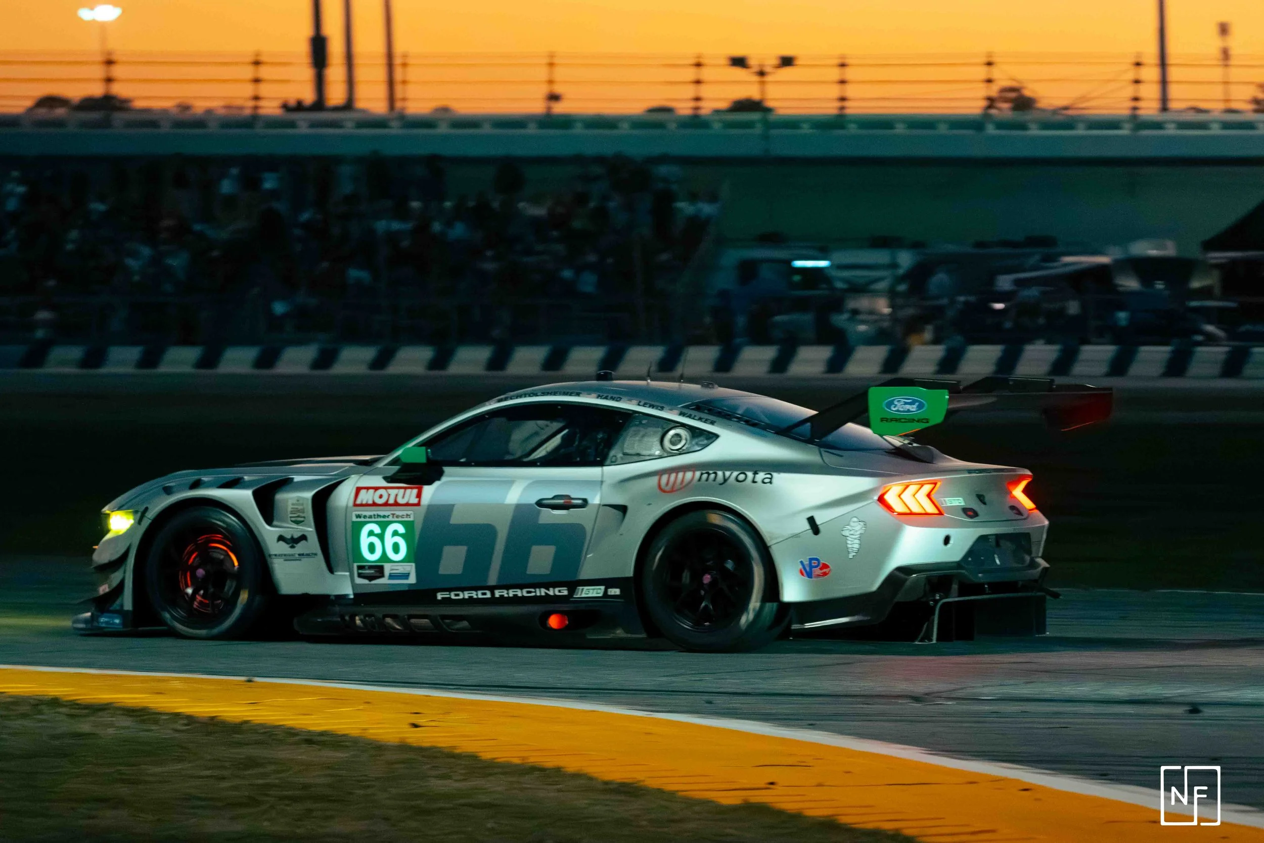 A race car on a race track during sunset, with a blurred crowd in the background. The car is silver with various sponsor logos, the number 66, and a rear wing with Ford branding.
