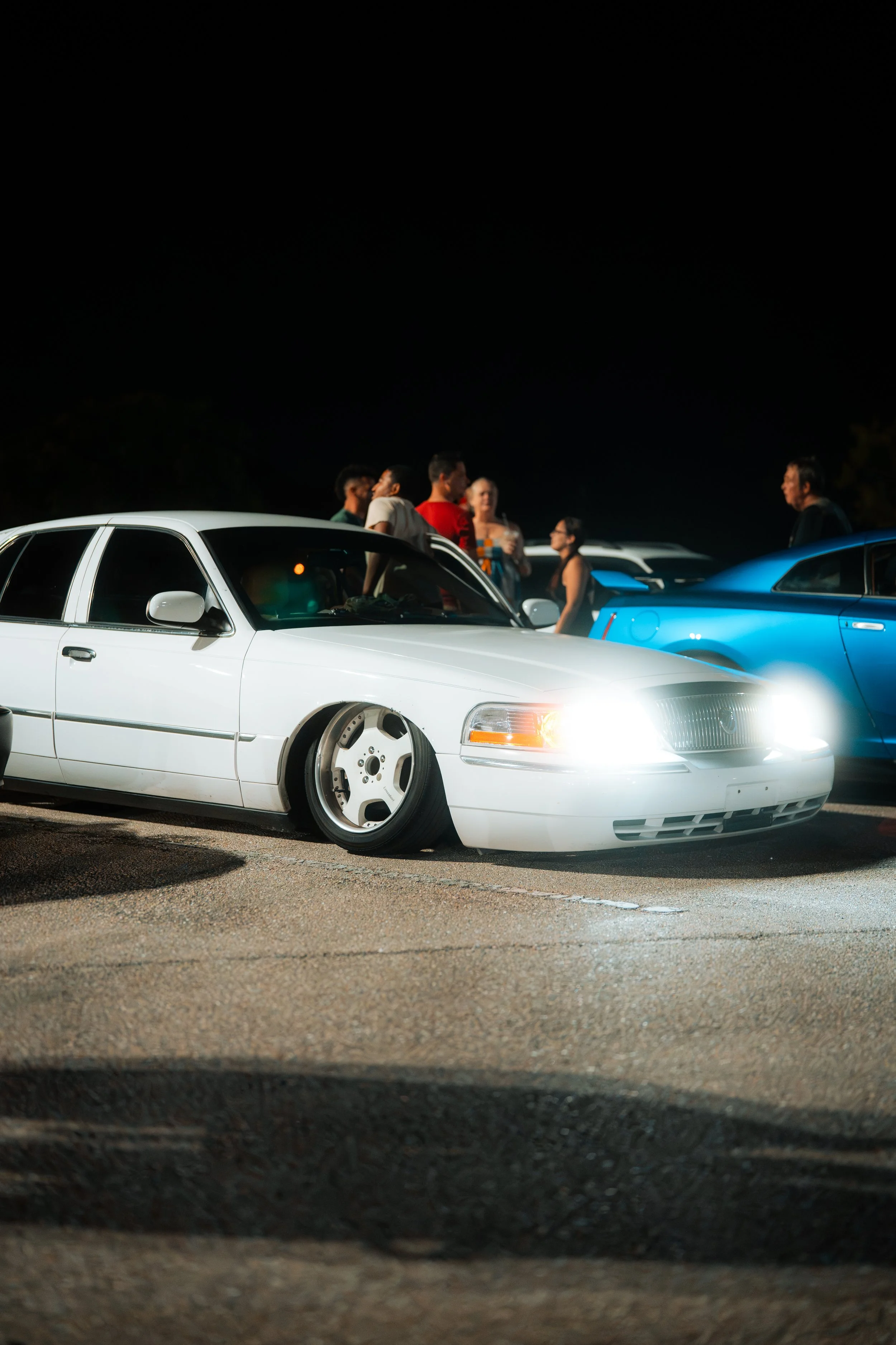 Nighttime car meet with a lowered white sedan with widened wheels and bright headlights, and a group of people in the background.