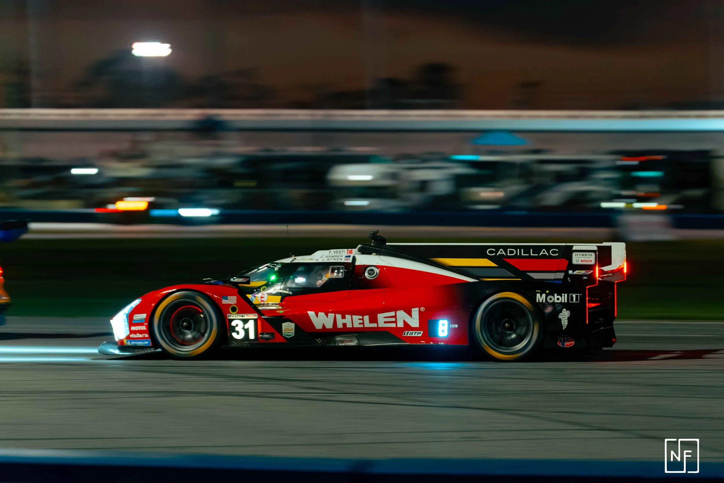 A red and black race car with the number 31 on its side driving on a racetrack at night, with blurred background lights.
