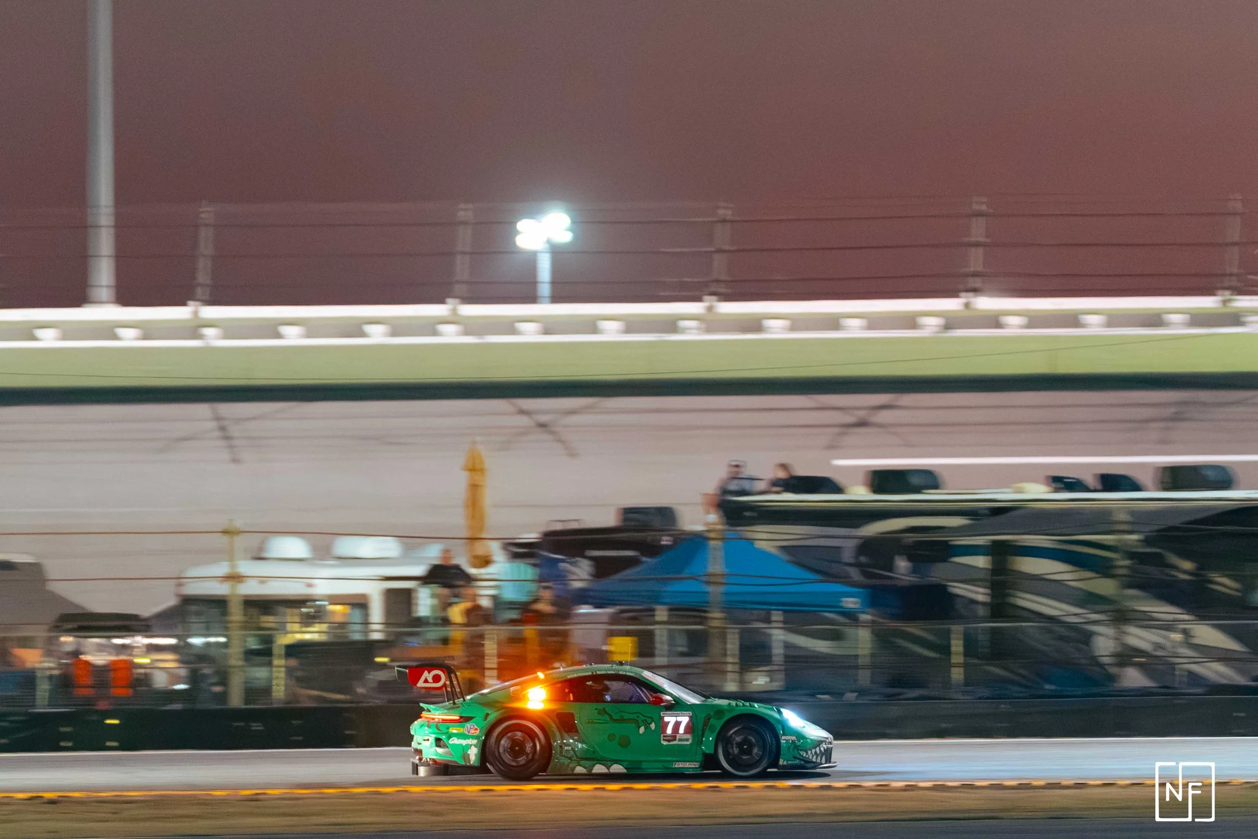 A green race car with the number 77 on its side driving on a race track at night, with a blurred background of tents, RVs, and grandstands.
