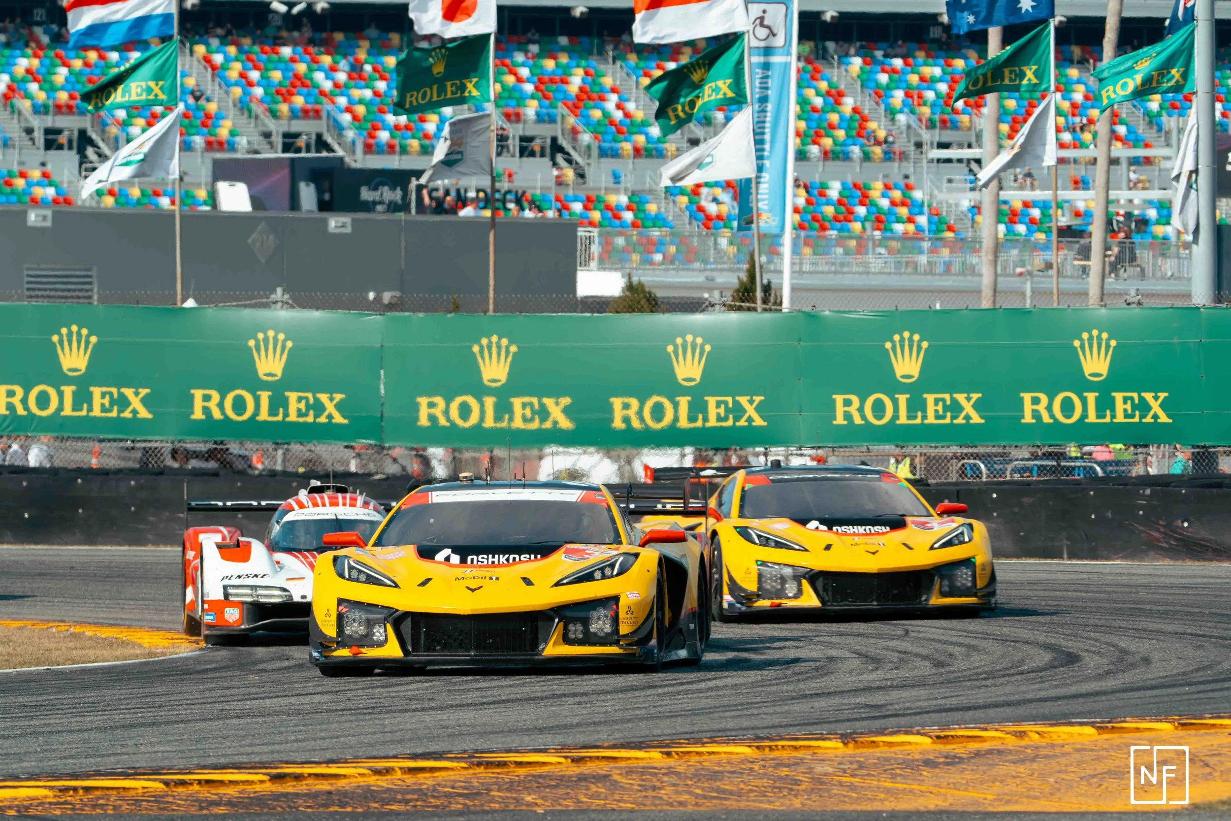 Two yellow race cars and one white race car on a racing track during a motorsport event, with green Rolex banners and colorful grandstands in the background.