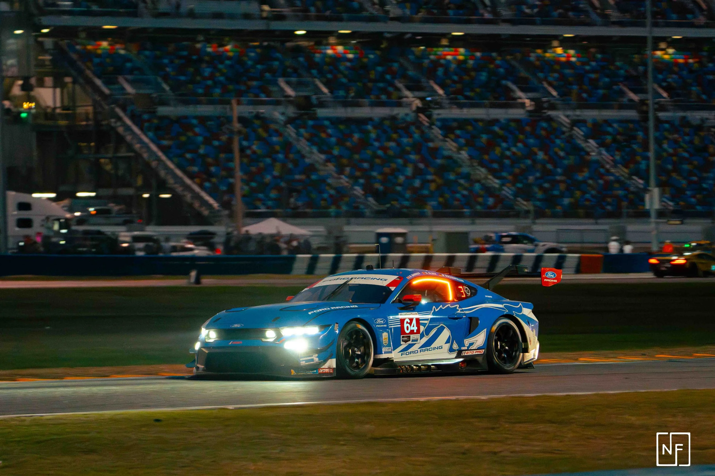 A blue Ford race car with number 64 on the side, racing on a track during nighttime, with illuminated headlights and a grandstand filled with colorful seats in the background.