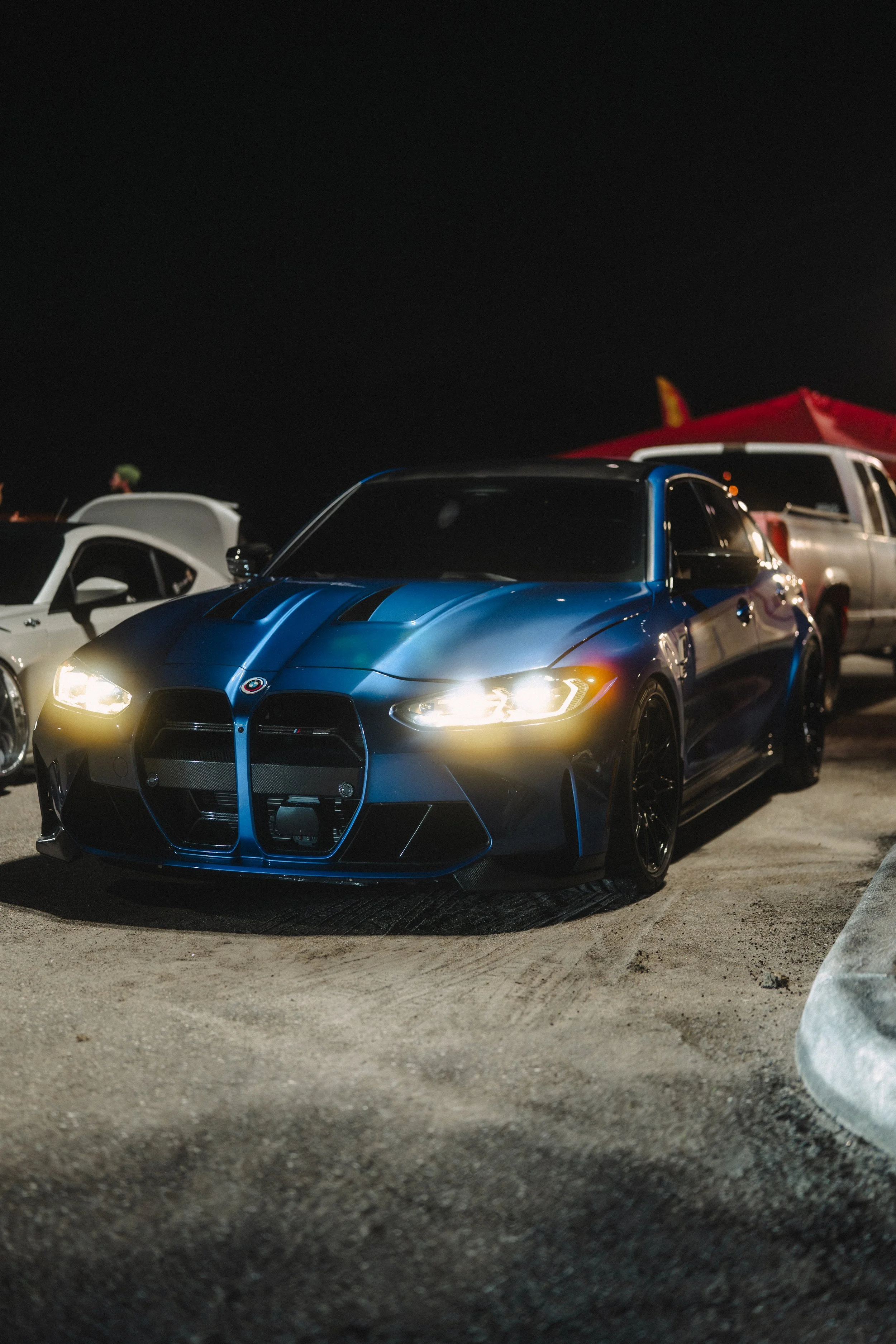 A dark blue sports car with distinctive front grille headlights glowing, parked at night, surrounded by other vehicles and a red canopy tent.