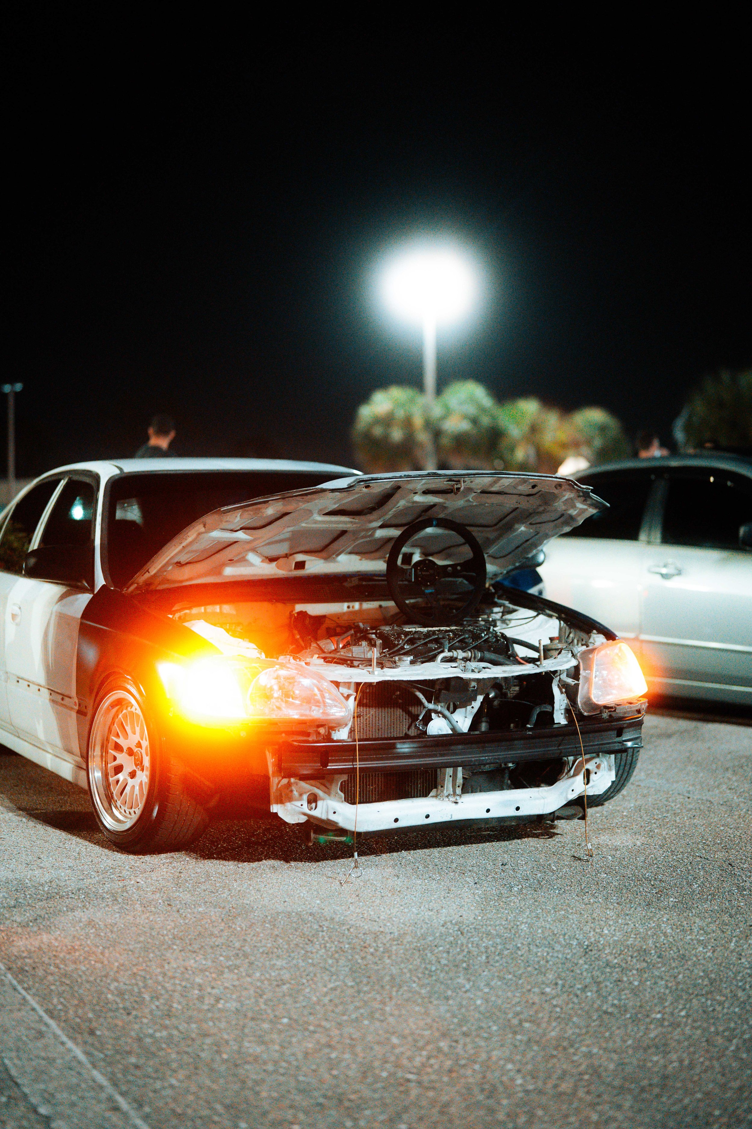 A damaged sports car with an open hood, parked at night in a parking lot, with its front bumper and grille missing and wires hanging. The car's left front signal light is on, and there are other cars and people in the background under a bright street